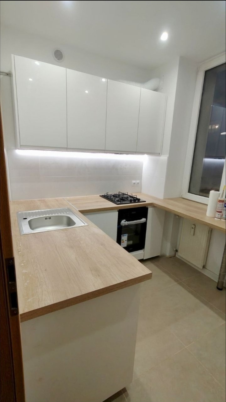 Nowoczesna, jasna kuchnia z białymi, błyszczącymi szafkami wiszącymi, blatem imitującym drewno i wbudowaną płytą gazową. Nad blatem zamontowane oświetlenie LED. Widoczny fragment zlewu i okna.