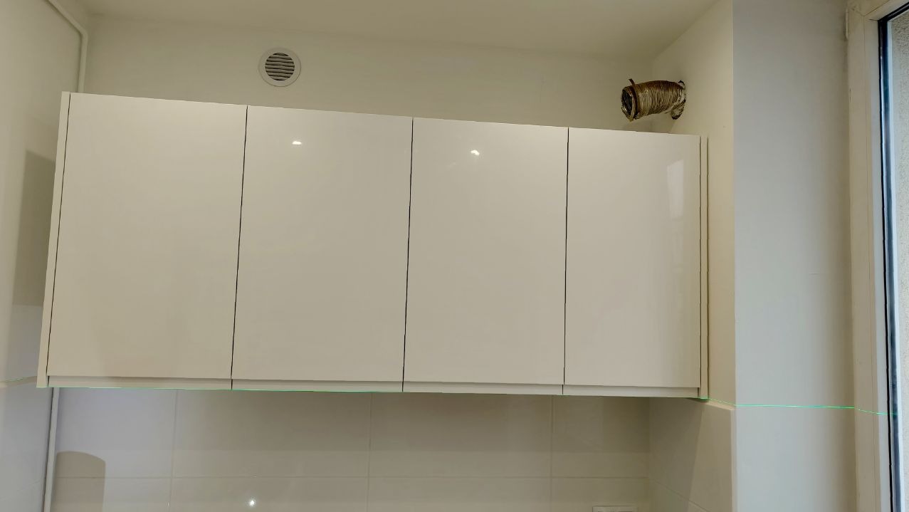 Białe, błyszczące szafki kuchenne wiszące nad płytkami, z widoczną rurą wentylacyjną na ścianie i zieloną linią lasera poziomującego.