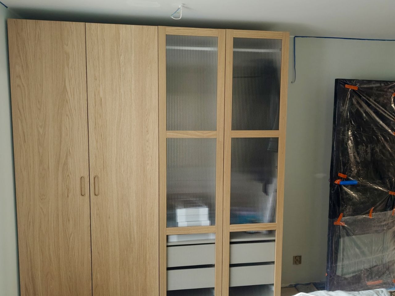 Jasny, drewniany, trzydrzwiowy segment szafy z ryflowanymi szybami w dwóch drzwiach, widoczne białe szuflady, obok drzwi zabezpieczone folią malarską.