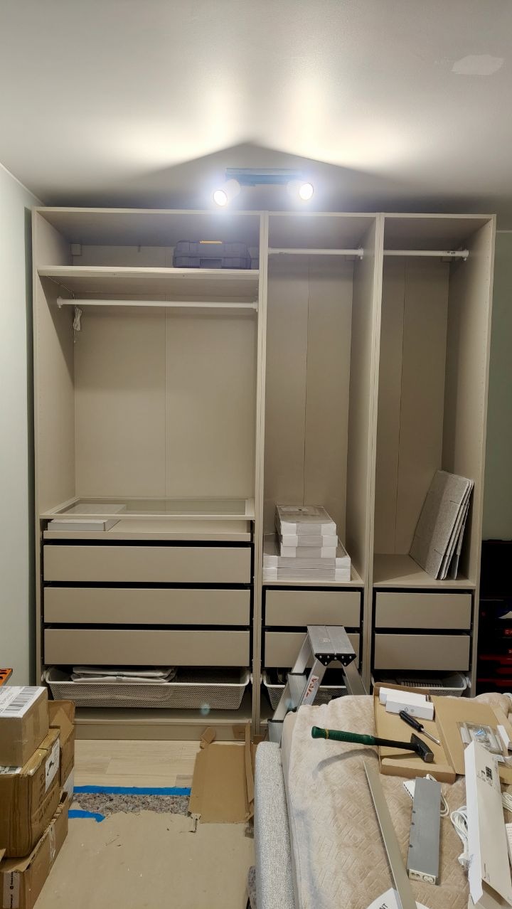 Wnętrze szafy z szufladami i półkami, częściowo zamontowanej, widoczne narzędzia pracy i kartony na podłodze, lampa oświetlająca wnętrze szafy.