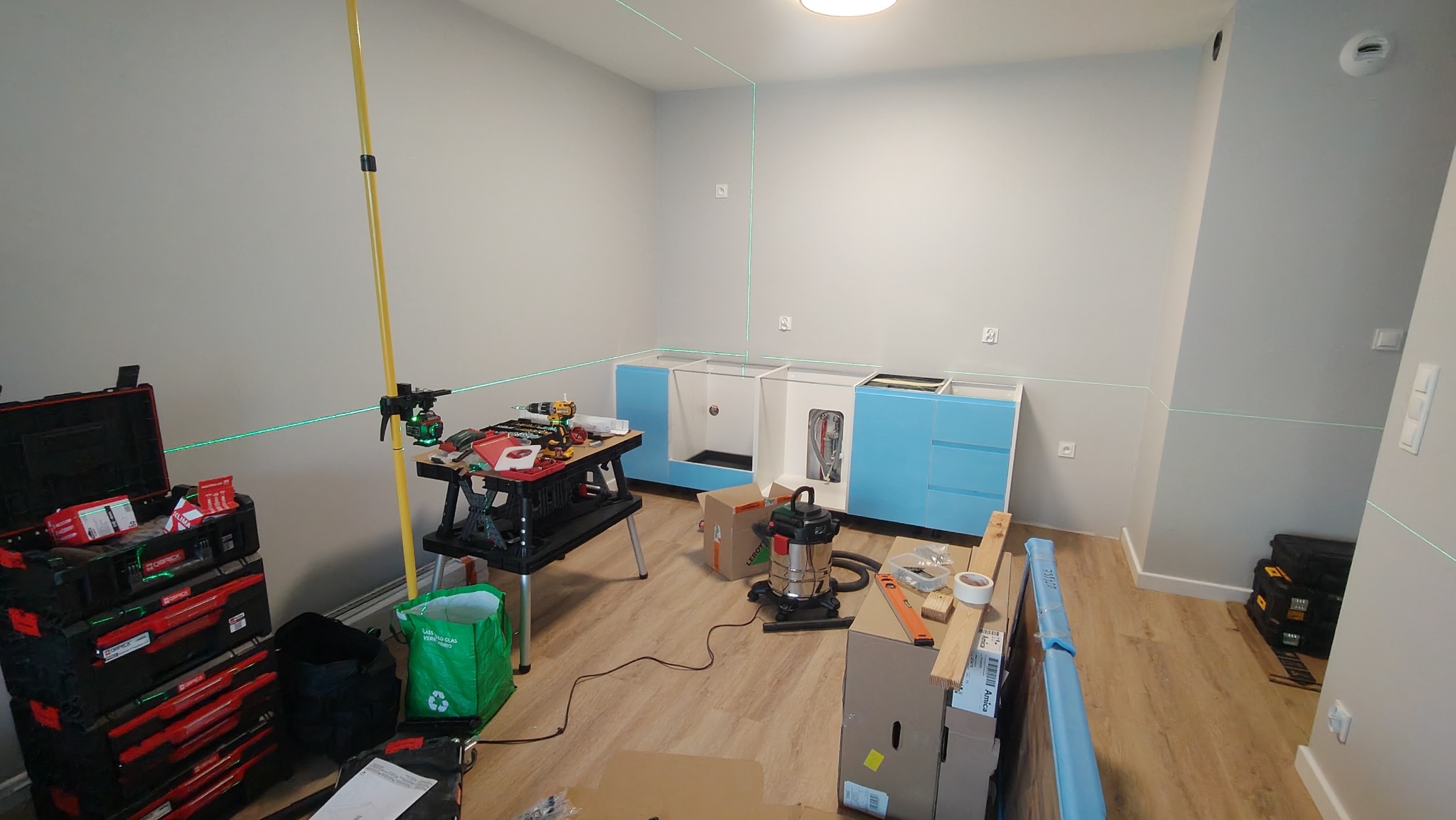Wnętrze w trakcie montażu mebli kuchennych: niebieskie szafki dolne ustawione wzdłuż ściany, widoczny laser krzyżowy wyznaczający poziom, narzędzia i akcesoria rozłożone na podłodze i stole...