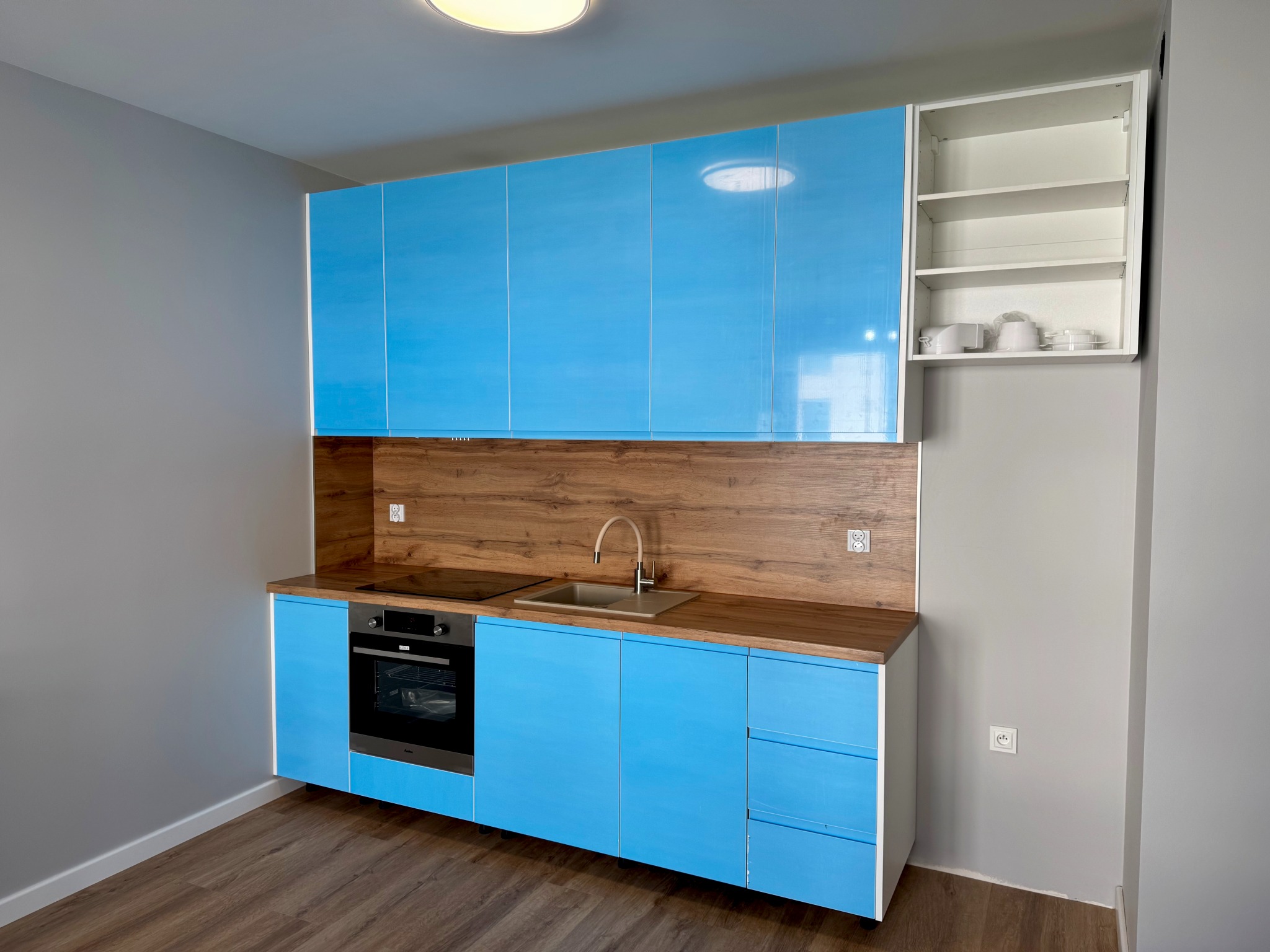 Niebieska, błyszcząca zabudowa kuchenna z drewnianym blatem i panelem, uzupełniona o białą szafkę wiszącą. Widok na kompletną, nową kuchnię w szarym pomieszczeniu.