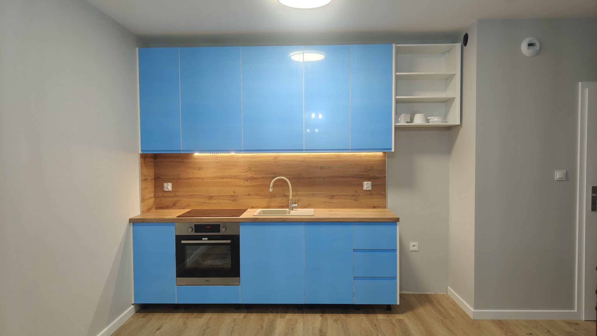 Błękitna, nowoczesna kuchnia z drewnianym blatem i oświetleniem podszafkowym. Widoczne białe półki z naczyniami i piekarnik w zabudowie. Całość w jasnym, minimalistycznym wnętrzu.