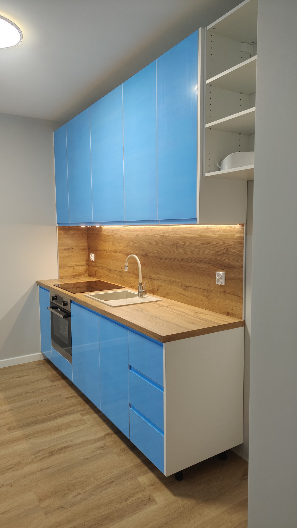 Błękitna, nowoczesna kuchnia z drewnianym blatem i oświetleniem podszafkowym. Widoczne szafki górne i dolne, piekarnik oraz zlew z baterią. Minimalistyczny styl.