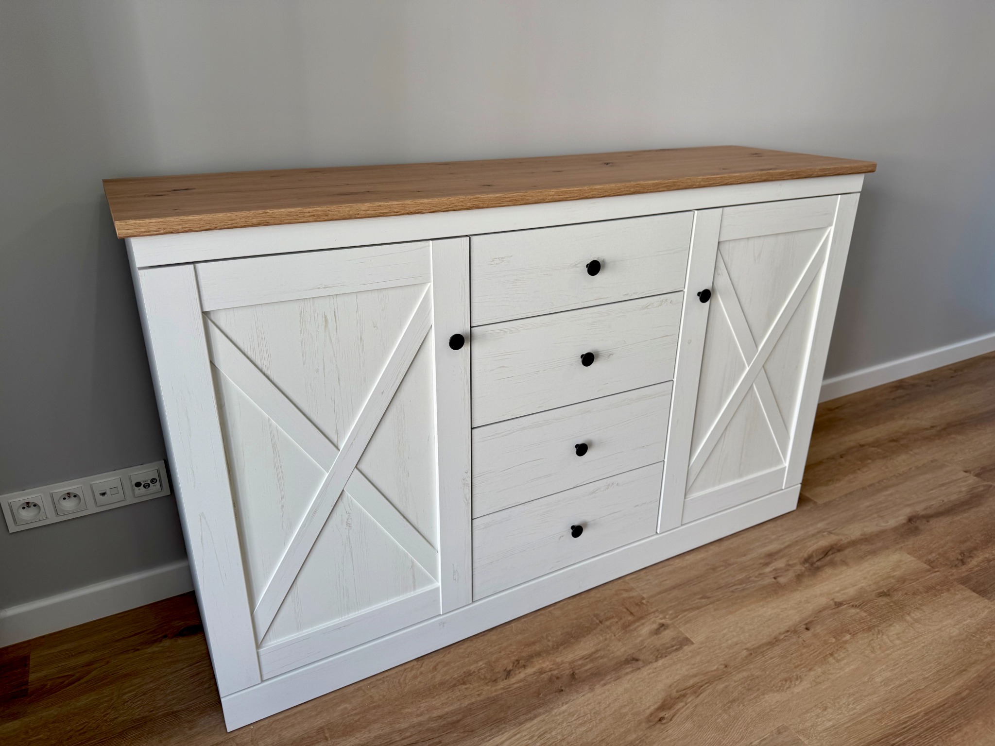 Biała komoda z drewnianym blatem i czarnymi uchwytami, stylizowana na rustykalną, stoi na panelach podłogowych przy szarej ścianie z gniazdkami.