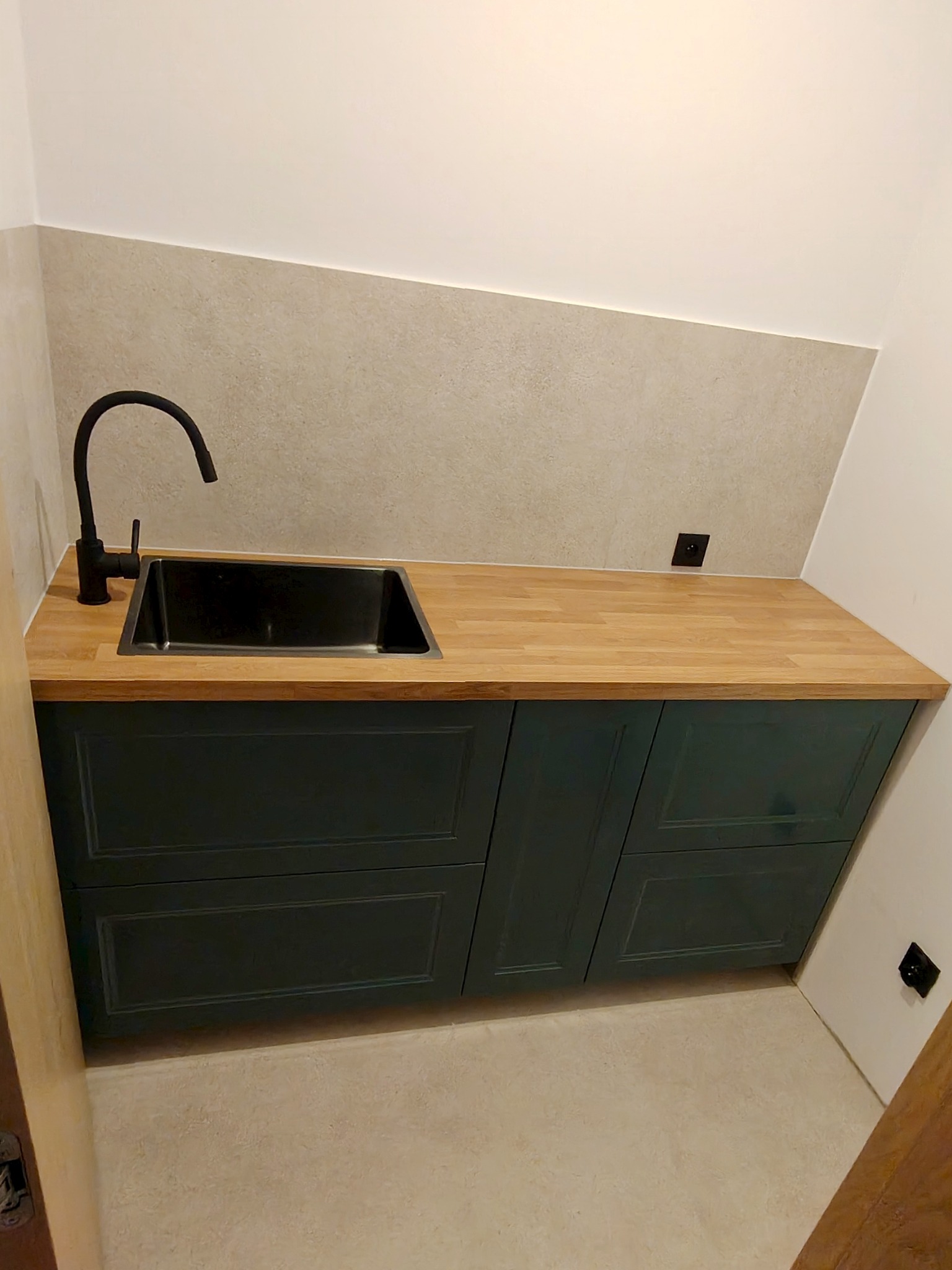 Nowoczesna szafka pod umywalkę z drewnianym blatem i czarną armaturą, zamontowana w łazience z beżową glazurą. Widoczne gniazdka elektryczne.