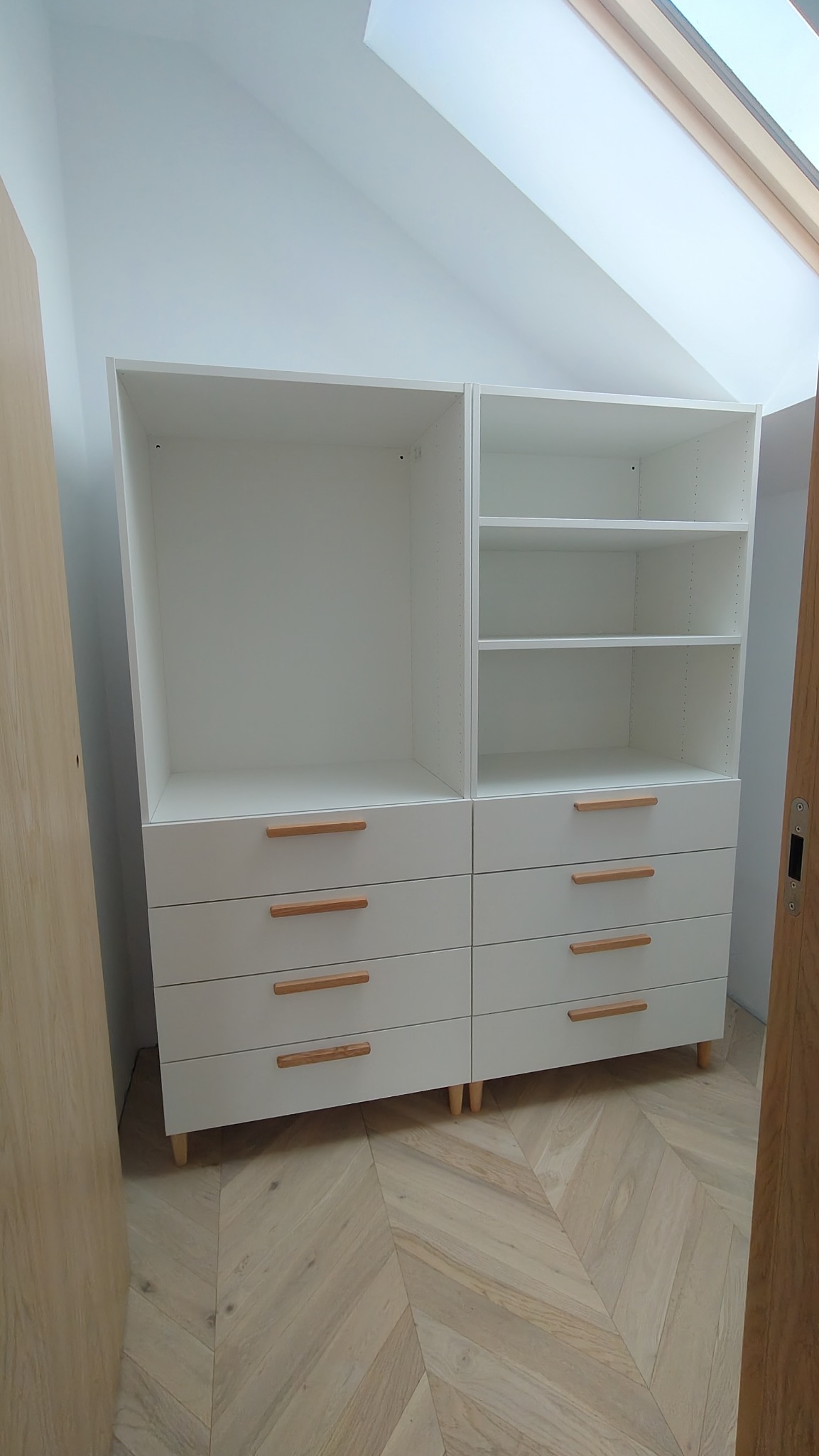 Biała szafa z szufladami i półkami, drewniane uchwyty, na parkiecie w jodełkę, w pokoju ze skosem i oknem dachowym. Częściowo widoczny fragment drzwi.