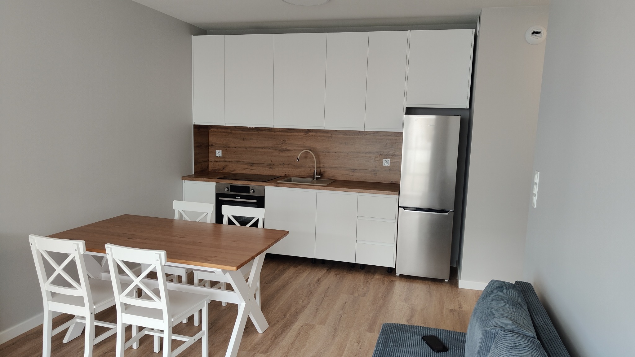 Nowoczesna kuchnia z białymi szafkami, drewnianym blatem i stołem z krzesłami. Uwagę przyciąga srebrna lodówka i drewnopodobny panel nad blatem.