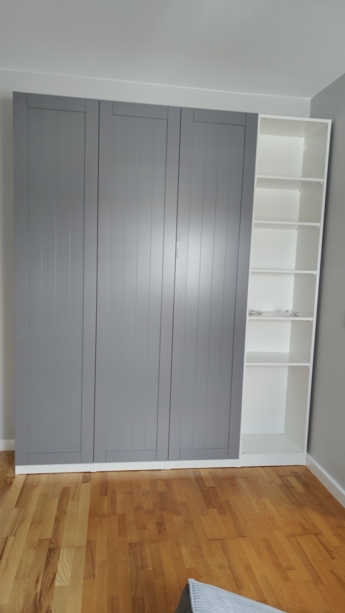 Szara szafa z trzema drzwiami i biały regał z półkami, zamontowane w rogu pokoju na drewnianej podłodze. Widok z dołu, oświetlenie naturalne.