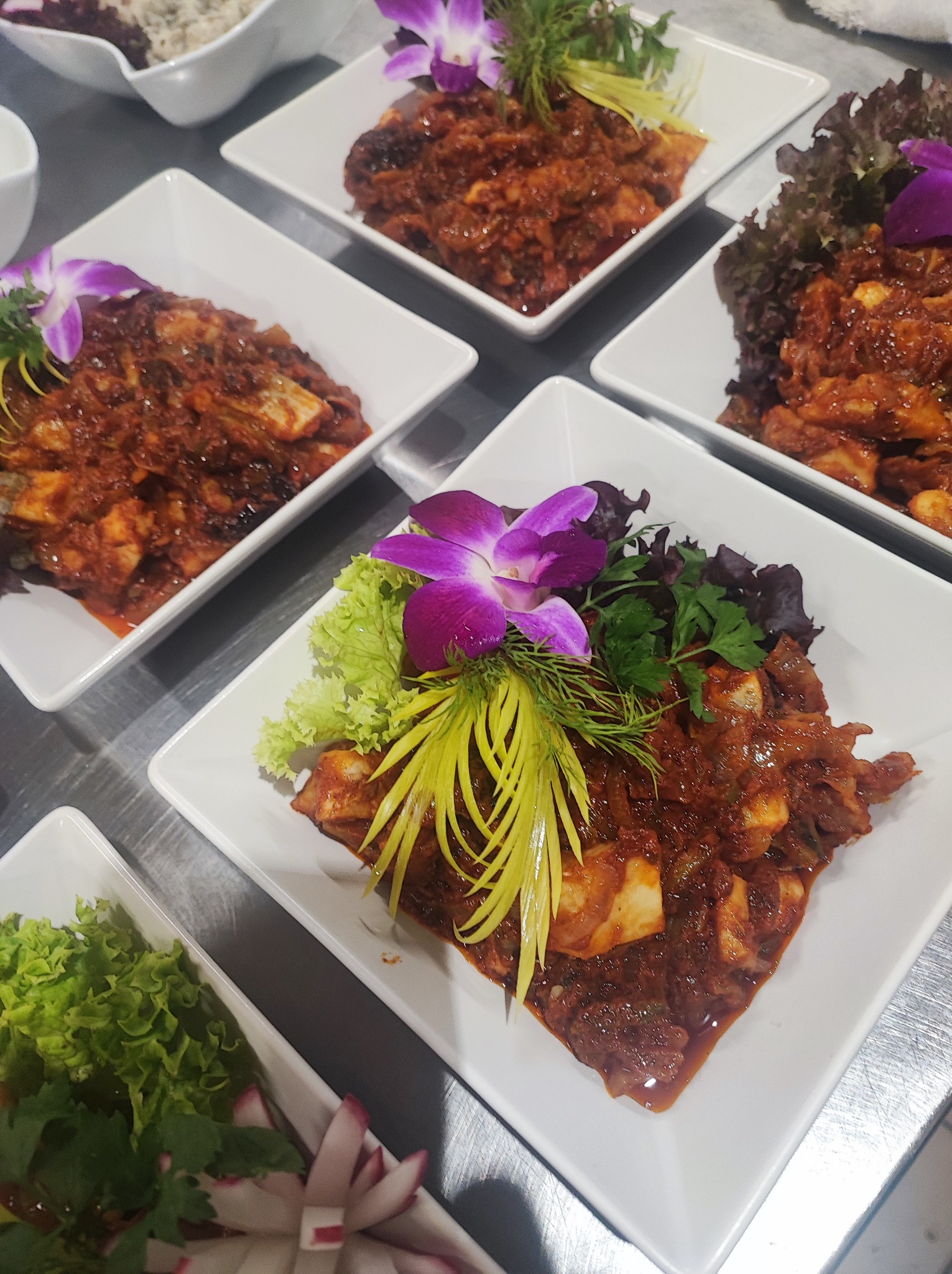 Elegancko podane dania mięsne w sosie na białych kwadratowych talerzach z dekoracją z orchidei, zieleniny i rzodkiewek, przygotowane do serwowania.