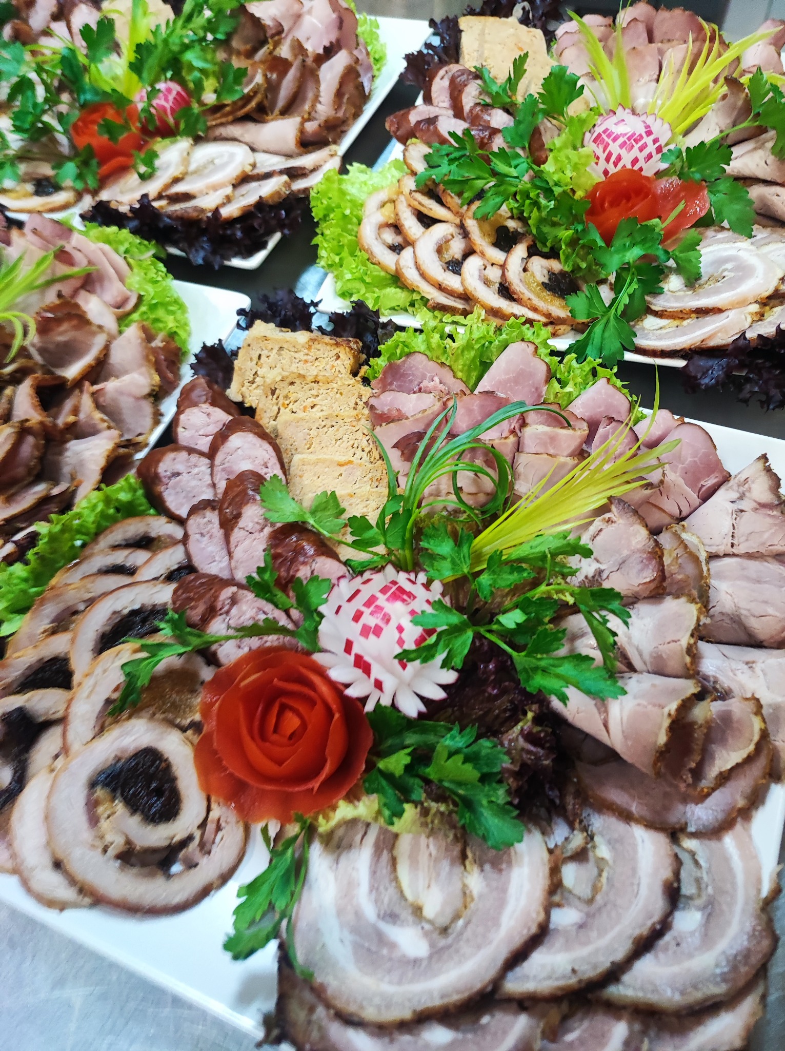 Dekoracyjnie ułożone wędliny i pasztety na białych paterach, ozdobione sałatą, natką pietruszki i rzeźbionymi rzodkiewkami.