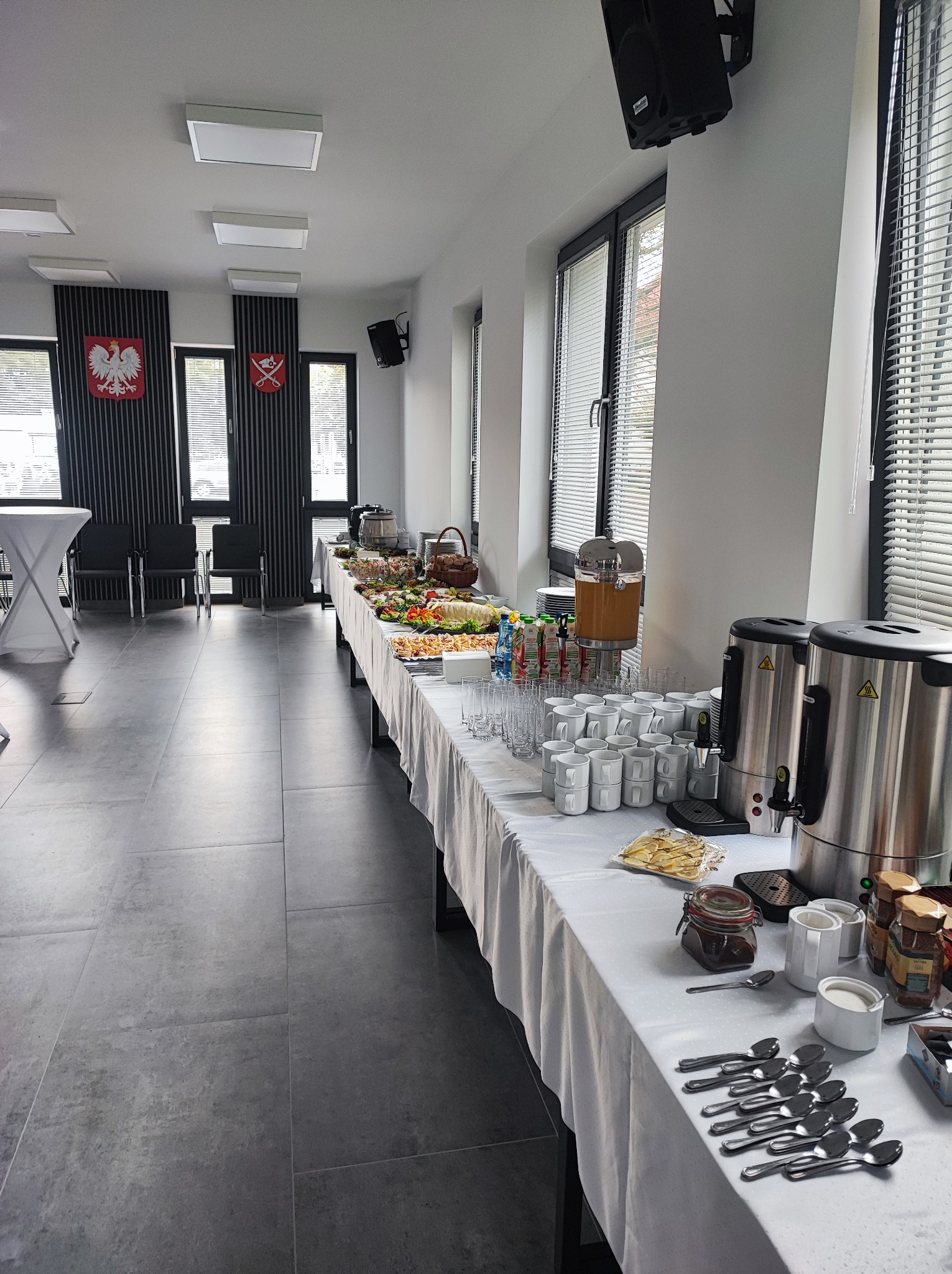 Długi stół z białym obrusem, zastawiony bufetem: sałatki, przekąski, napoje, ekspresy do kawy, filiżanki i sztućce w jasnym pomieszczeniu z oknami w żaluzjach i flagami państwowymi.