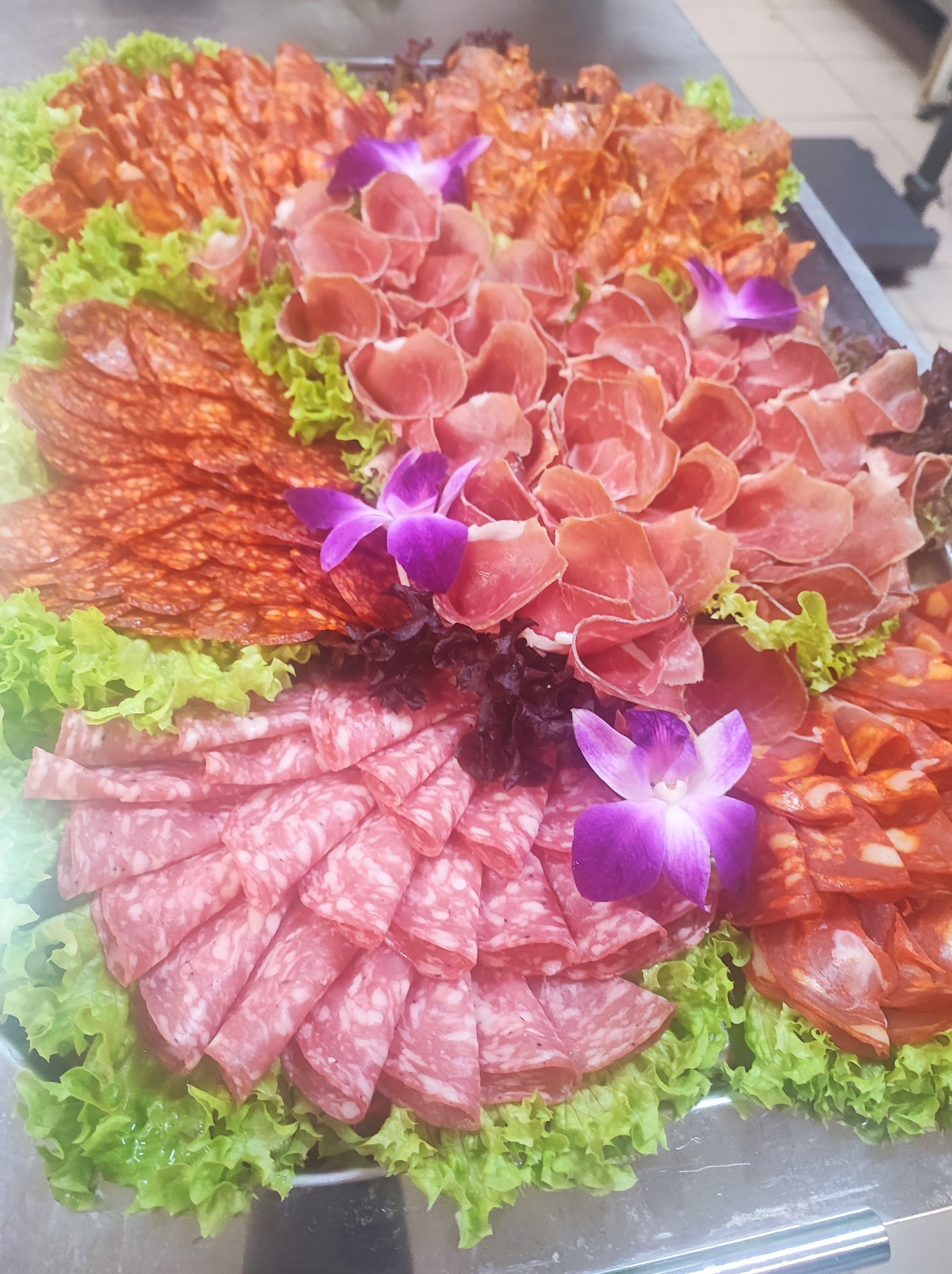 Dekoracyjna taca z różnymi rodzajami wędlin, salami, szynki i chorizo, ułożonymi na świeżej sałacie i ozdobionymi fioletowymi kwiatami.