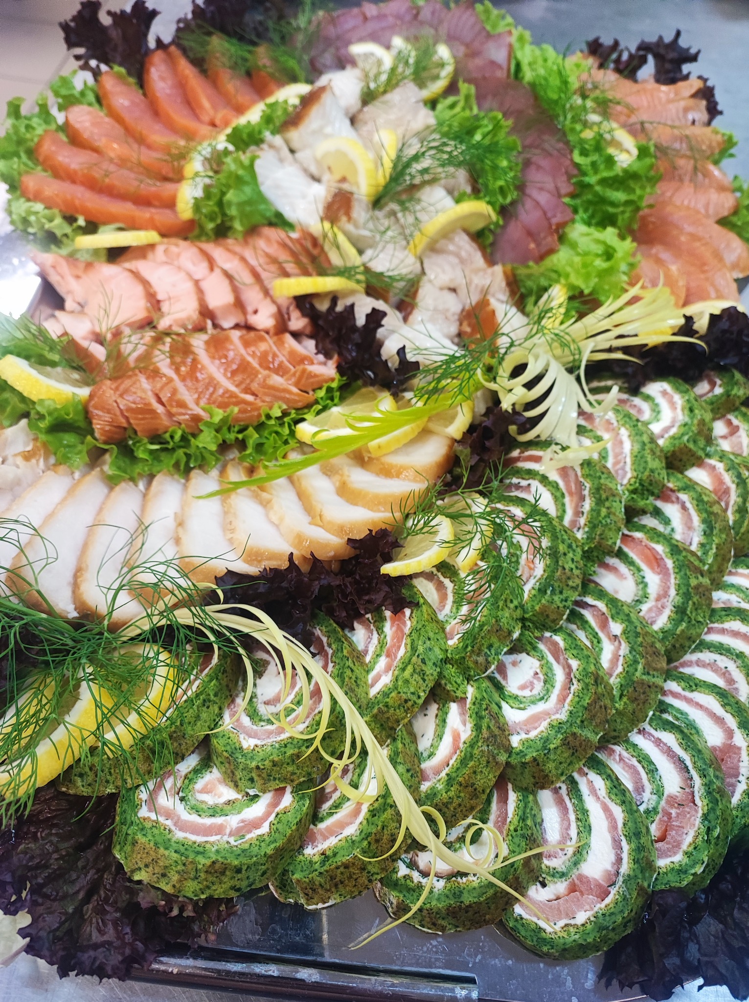 Dekoracyjna taca z różnorodnymi przystawkami rybnymi: roladki ze szpinaku i łososia, wędzone ryby w plastrach, plastry łososia, dekorowane sałatą, koperkiem i cytryną.