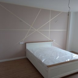 Nazar Semkulych - Pudrowo-różowa ściana z geometrycznym wzorem z taśmy malarskiej nad białym łóżkiem z drewnianymi akcentami, widoczne gniazdka elektryczne i surowy kabel zwisający z sufitu.