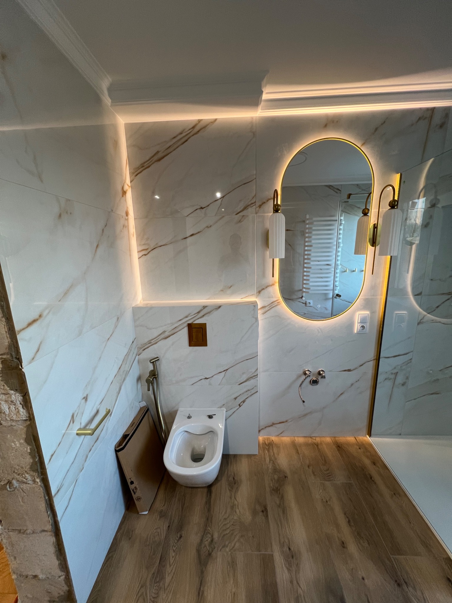 Nowoczesna łazienka z białymi marmurkowymi płytkami, podświetlanym lustrem owalnym i drewnopodobną podłogą, widoczny fragment ściany w trakcie prac wykończeniowych.