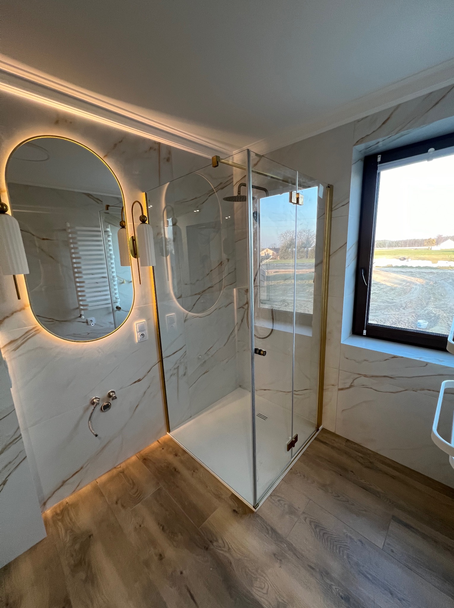 Nowoczesna łazienka z kabiną prysznicową z pozłacanymi detalami, podświetlanym lustrem owalnym i płytkami imitującymi marmur, z widokiem na pole przez okno.