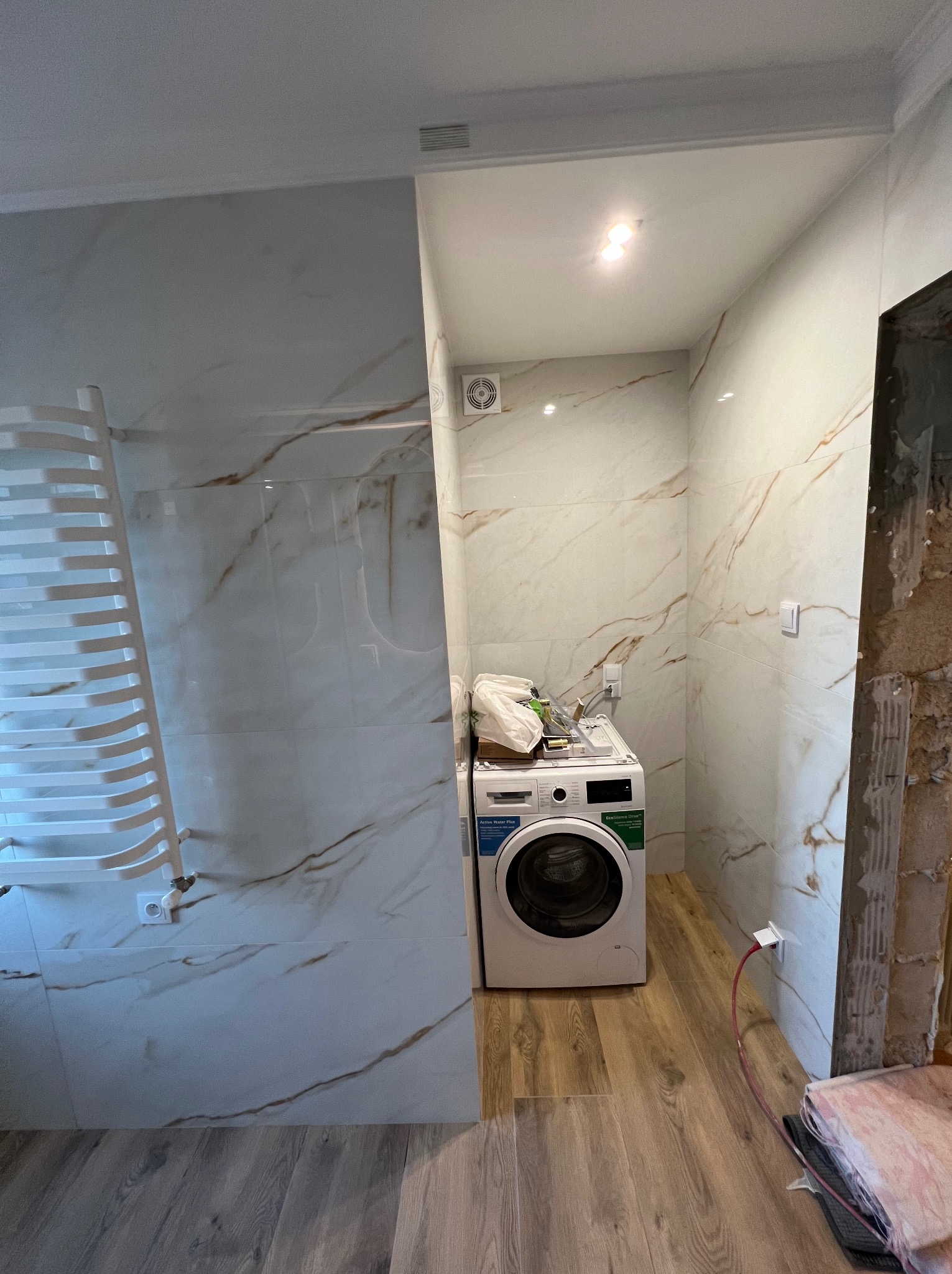 Wnętrze łazienki w trakcie remontu w Piasecznie, widoczne płytki imitujące marmur na ścianach, grzejnik drabinkowy, pralka częściowo zasłonięta materiałami budowlanymi oraz fragment ściany w stanie...