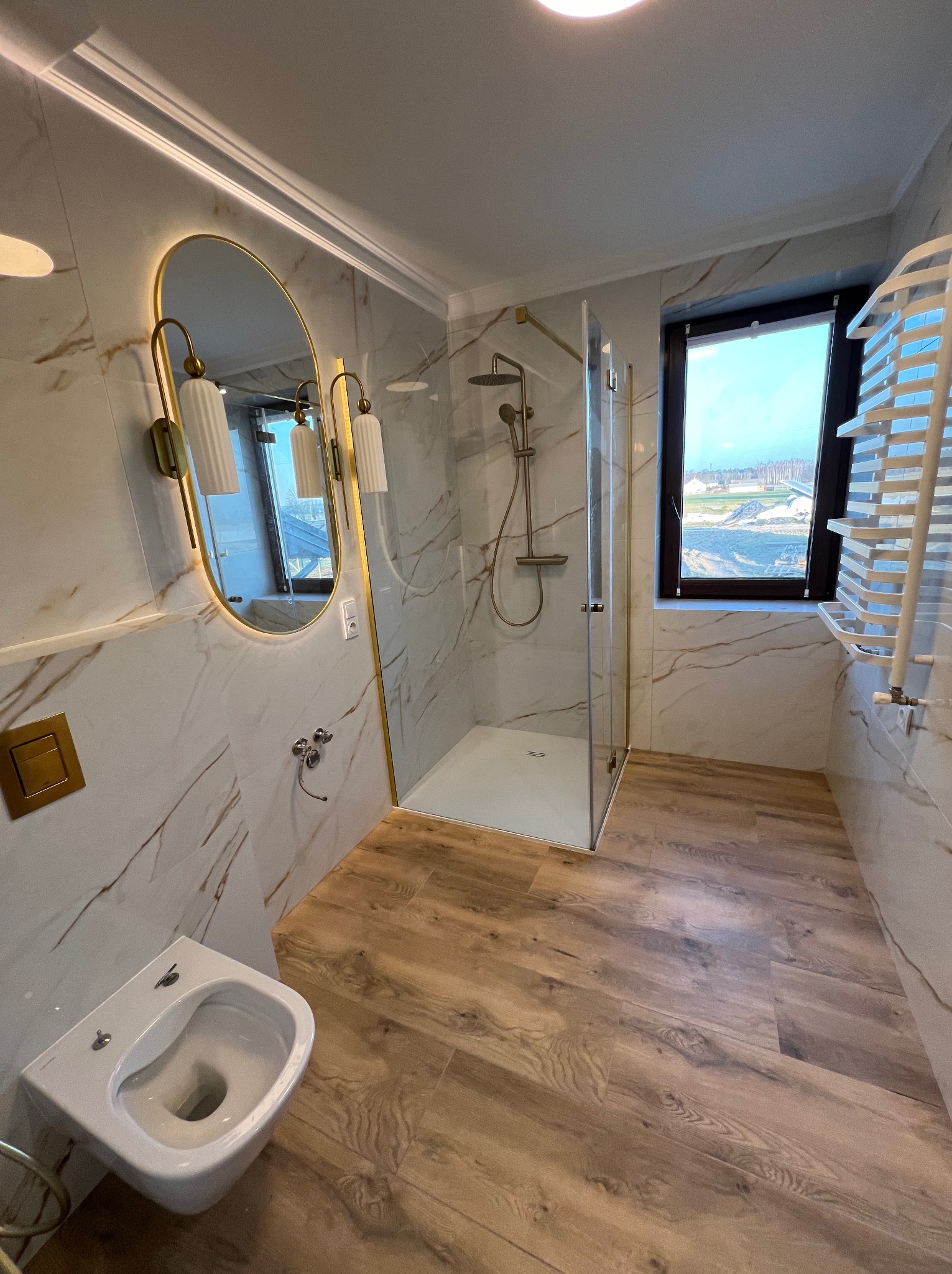 Nowoczesna łazienka z kabiną prysznicową ze złotymi detalami, owalnym lustrem z podświetleniem LED i marmurowymi płytkami na ścianach, drewnopodobna podłoga i widok z okna na teren budowy.