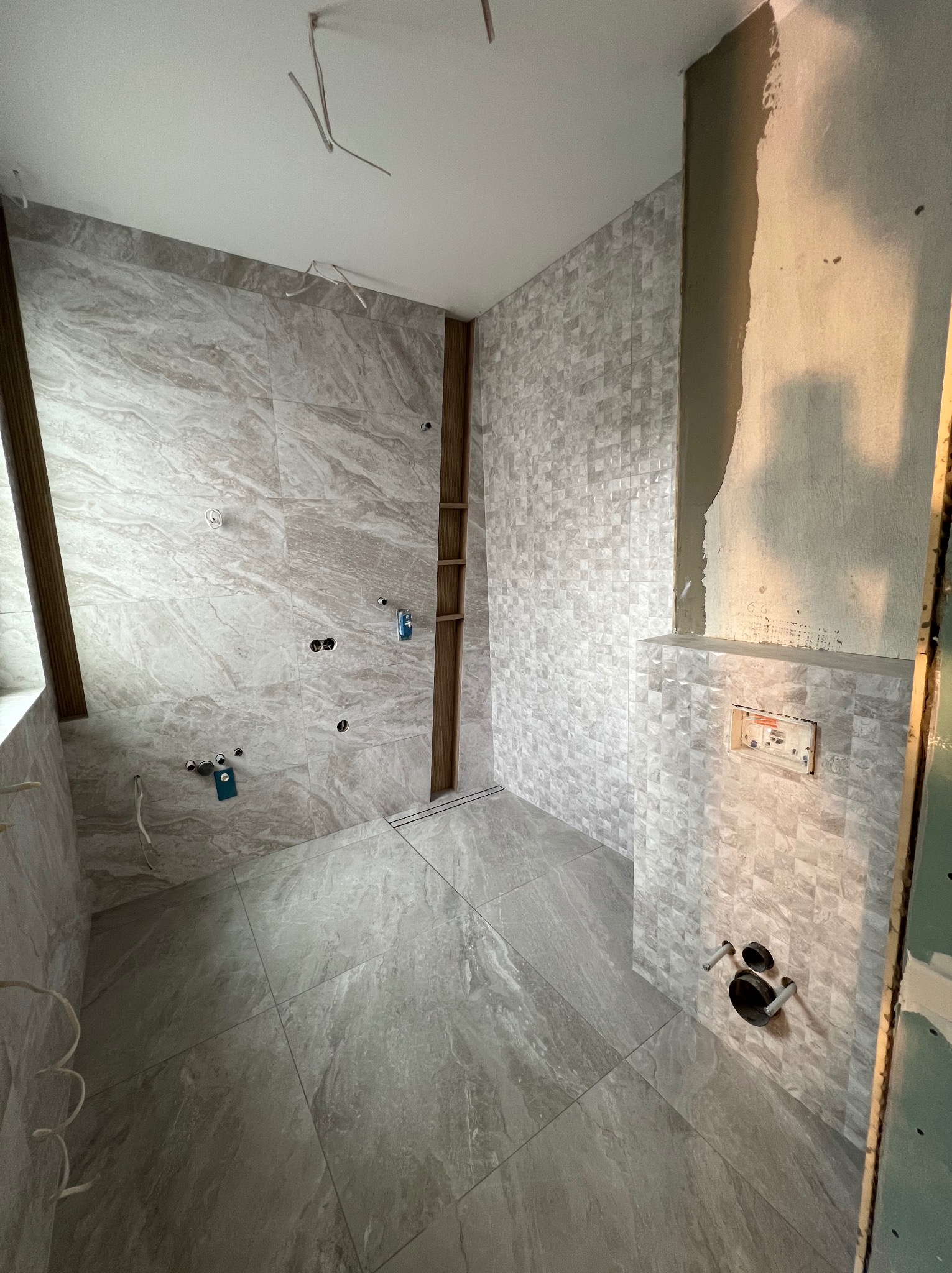 Wnętrze łazienki w trakcie remontu, widoczne położone płytki imitujące marmur na ścianach i podłodze, instalacja wodno-kanalizacyjna przygotowana do montażu armatury, surowa ściana z płyt...