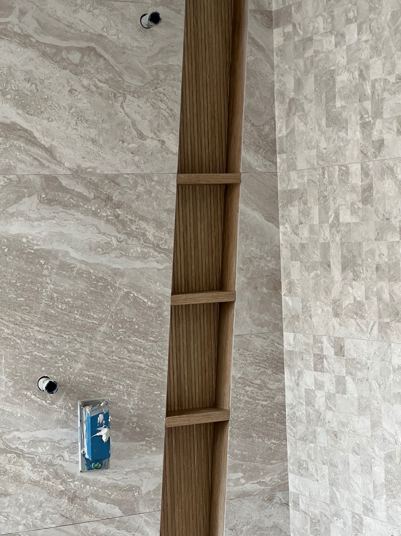 Montaż marmurowych płytek w łazience z drewnianą wnęką na półki, widoczne gniazdko elektryczne w trakcie instalacji.