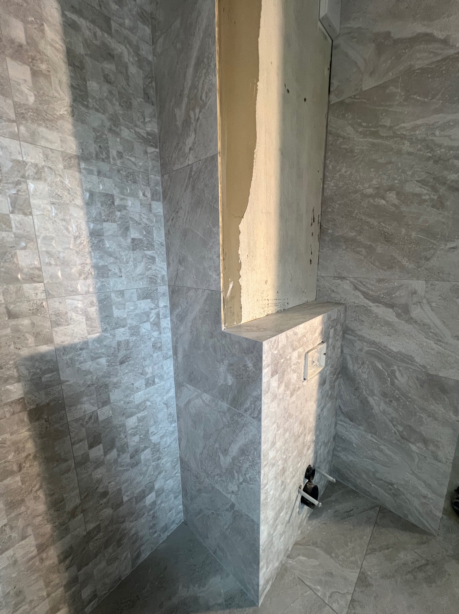 Fragment łazienki w trakcie remontu z szarymi płytkami imitującymi kamień, widoczne niedokończone obłożenie ściany i wystające rury instalacji wodnej.