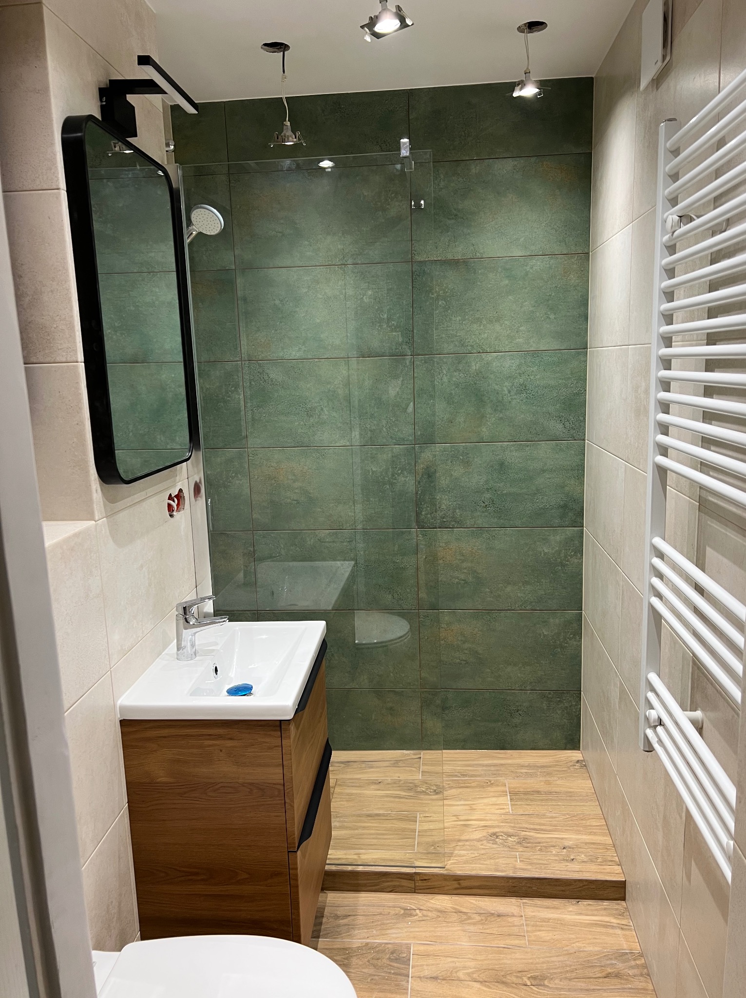 Nowoczesna łazienka z zielonymi płytkami w strefie prysznicowej, drewnopodobną podłogą i umywalką z szafką z ciemnego drewna, lustro w czarnej ramie.