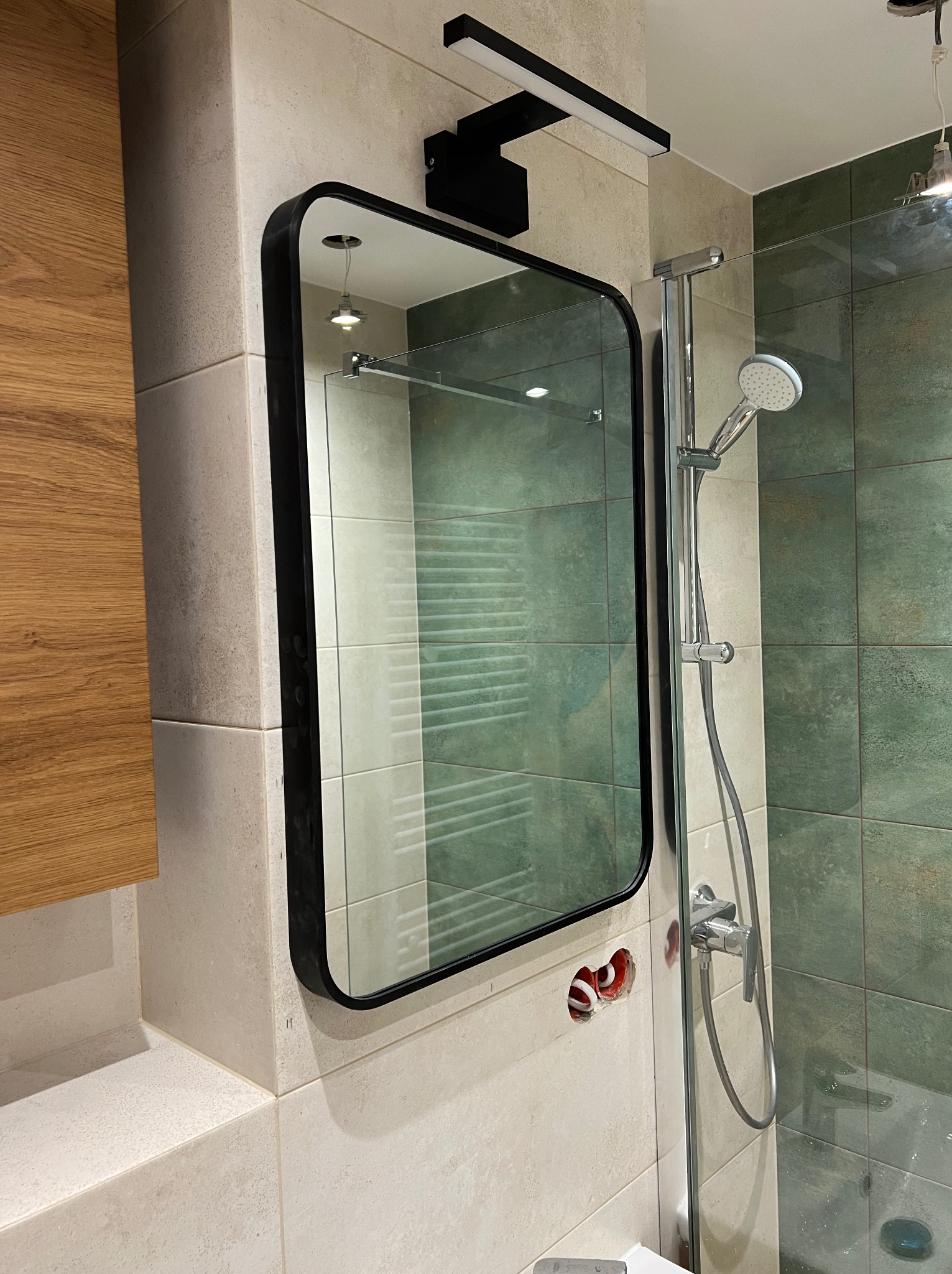 Nowoczesna łazienka w trakcie wykańczania: czarne, zaokrąglone lustro z podświetleniem LED, kabina prysznicowa ze srebrną armaturą, beżowe i zielone płytki, widoczne przewody elektryczne w ścianie.