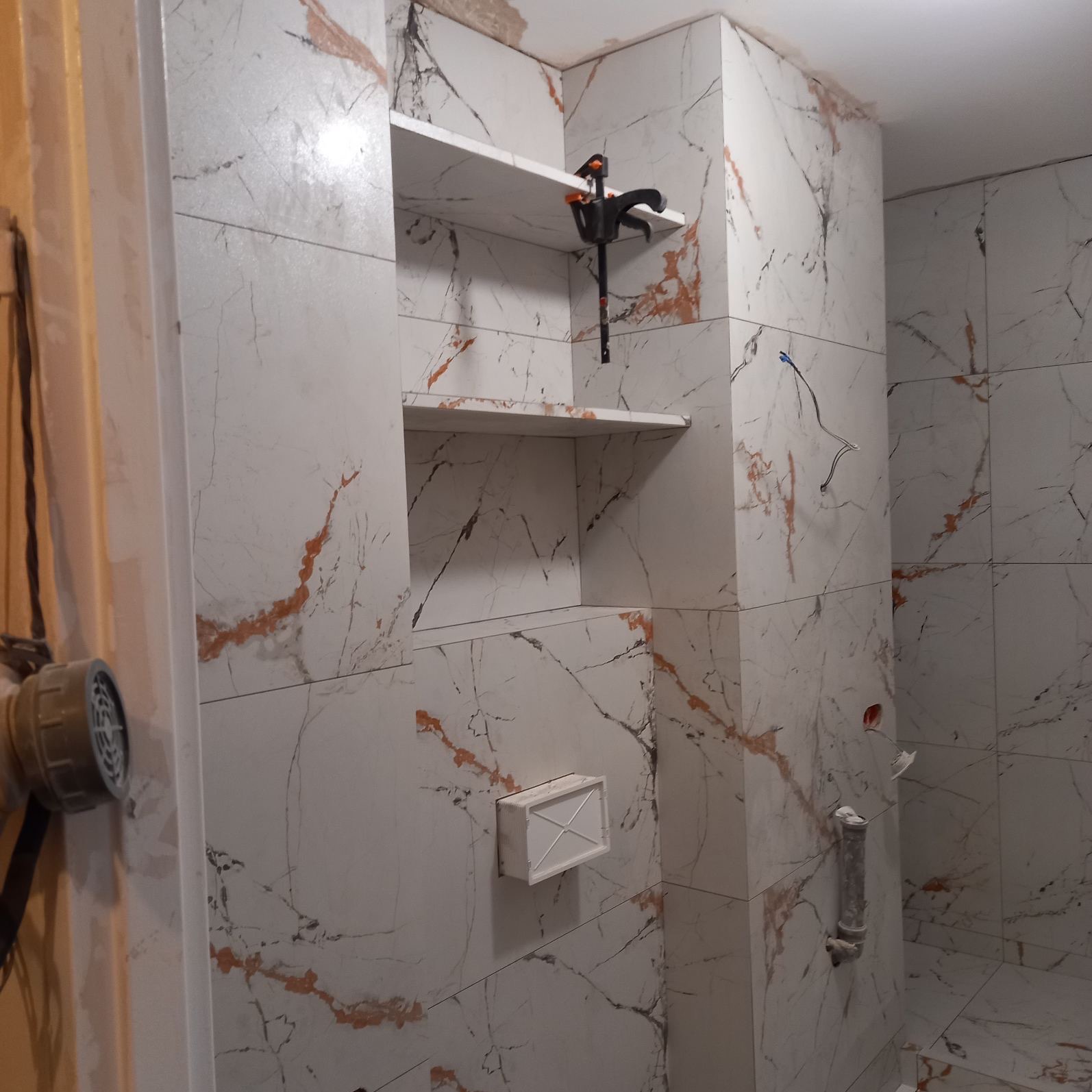 Ściana łazienki wyłożona białymi płytkami imitującymi marmur z wnękami półek, na jednej z półek zaciśnięty zacisk stolarski, widoczne wystające kable i rury kanalizacyjne.