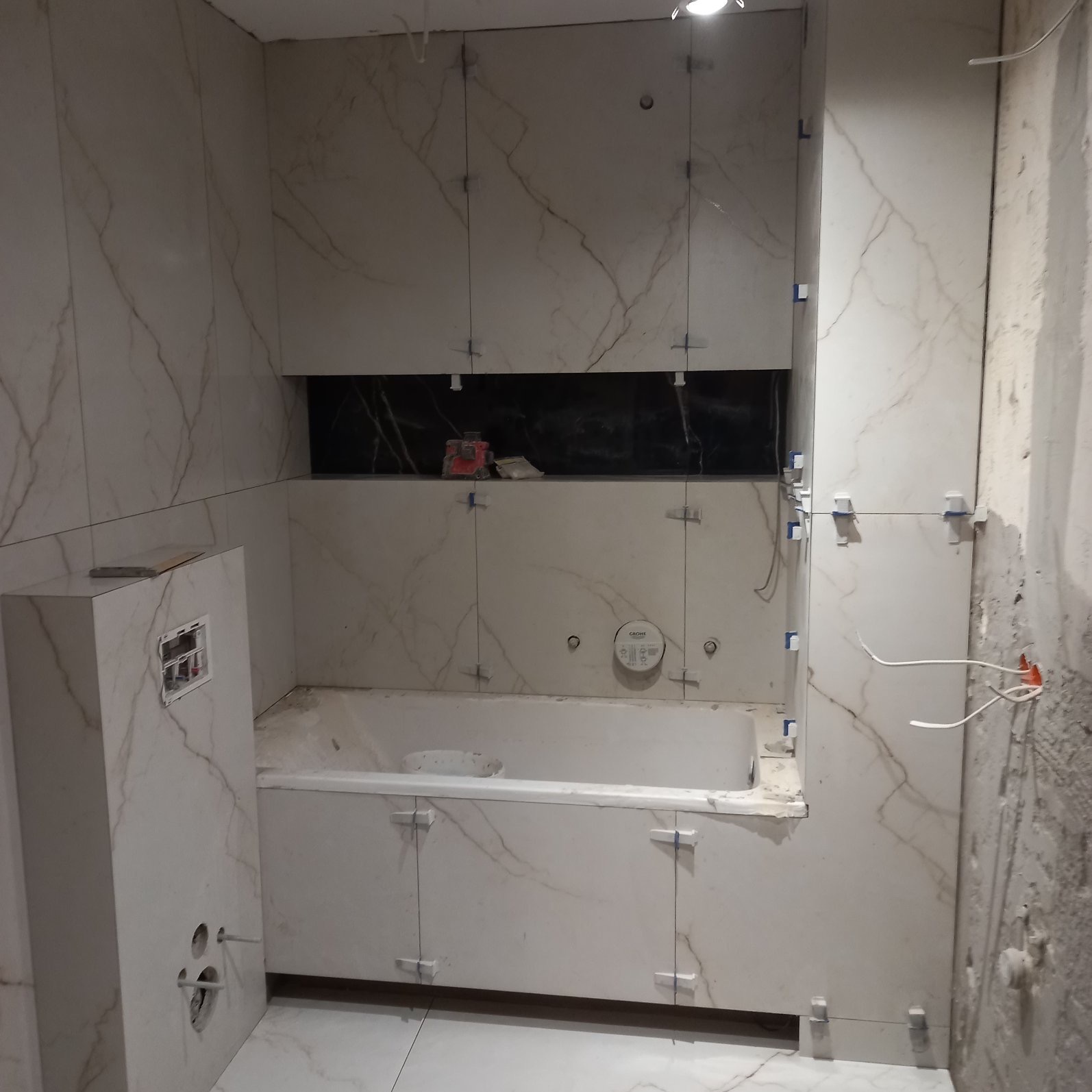 W trakcie prac: białe płytki imitujące marmur z ciemnymi żyłami, układane na ścianach i obudowie wanny, widoczne krzyżyki dystansowe, fragment instalacji wodnej Grohe, niezakończone prace...