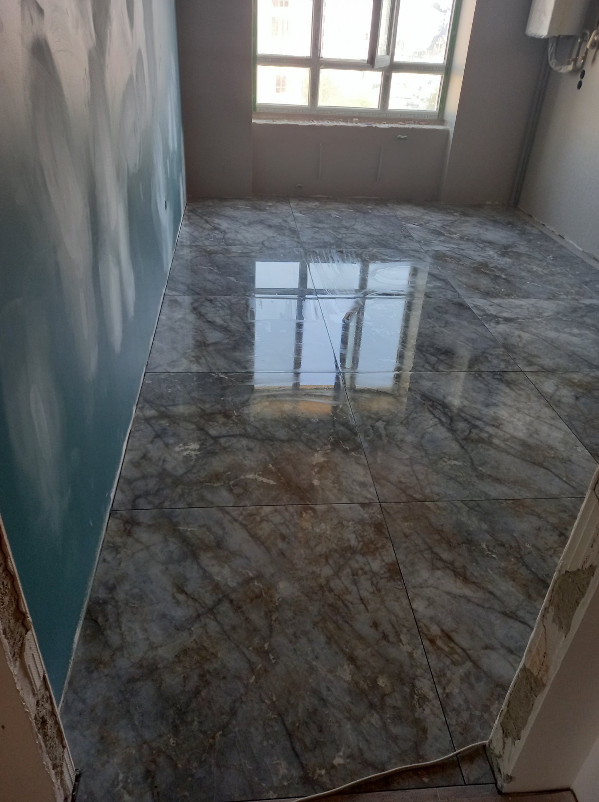 Wykończona podłoga z płytek imitujących marmur w jasnym pomieszczeniu z oknem, ściana częściowo przygotowana do malowania.