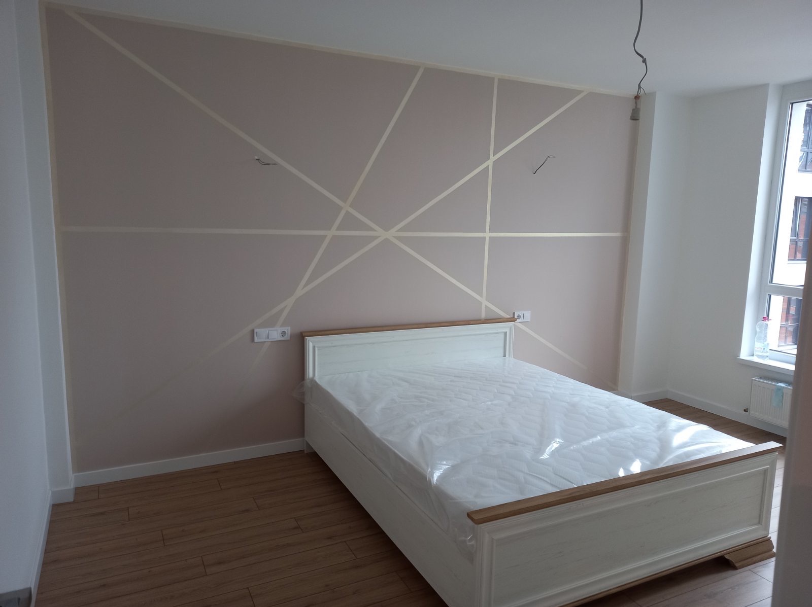 Pudrowo-różowa ściana z geometrycznym wzorem z taśmy malarskiej nad białym łóżkiem z drewnianymi akcentami, widoczne gniazdka elektryczne i surowy kabel zwisający z sufitu.