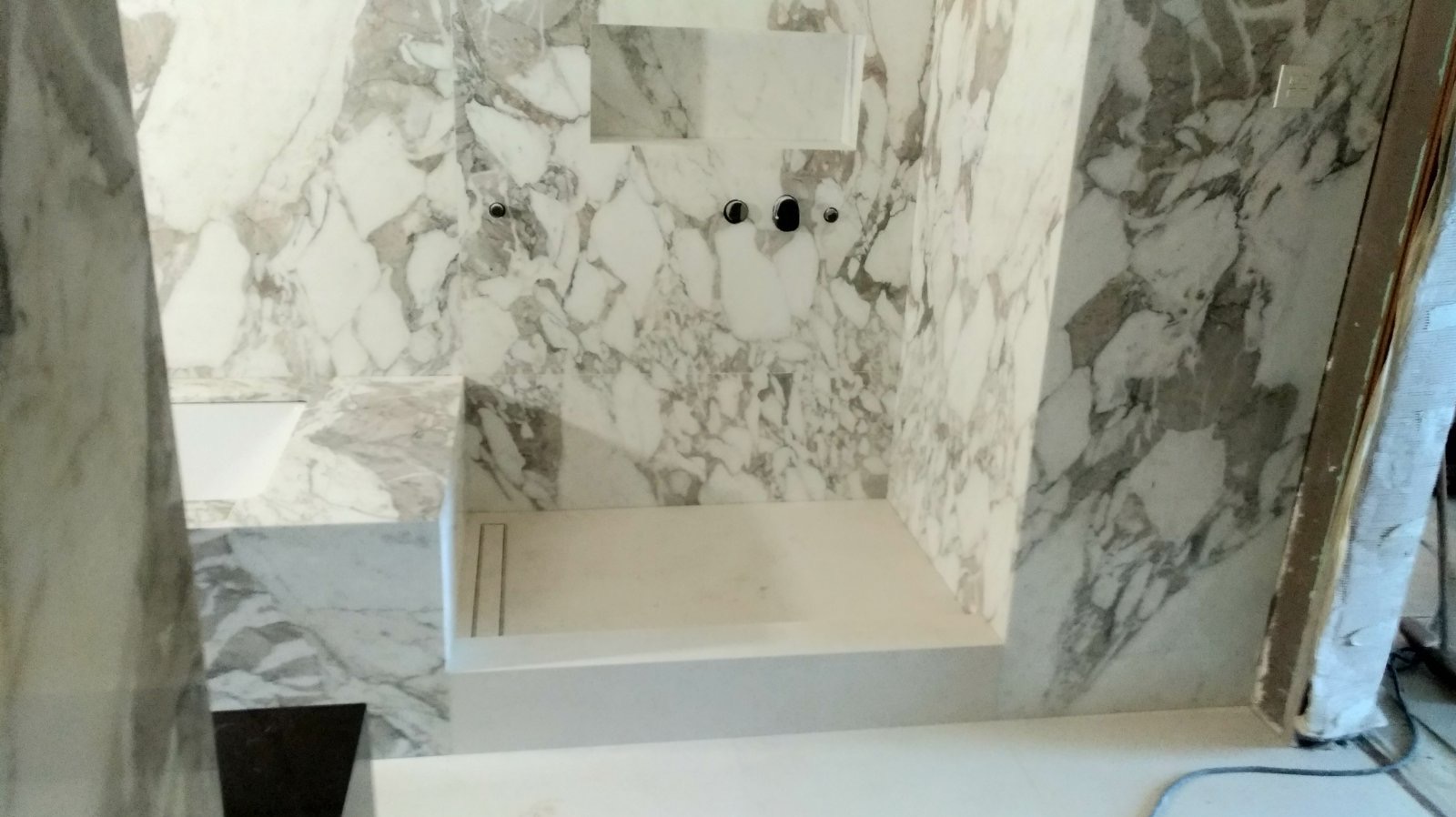 Wnętrze łazienki w trakcie prac wykończeniowych, ściany i obudowa wanny wyłożone marmurem z szarymi żyłami, widoczne czarne baterie oraz odpływ liniowy w brodziku.