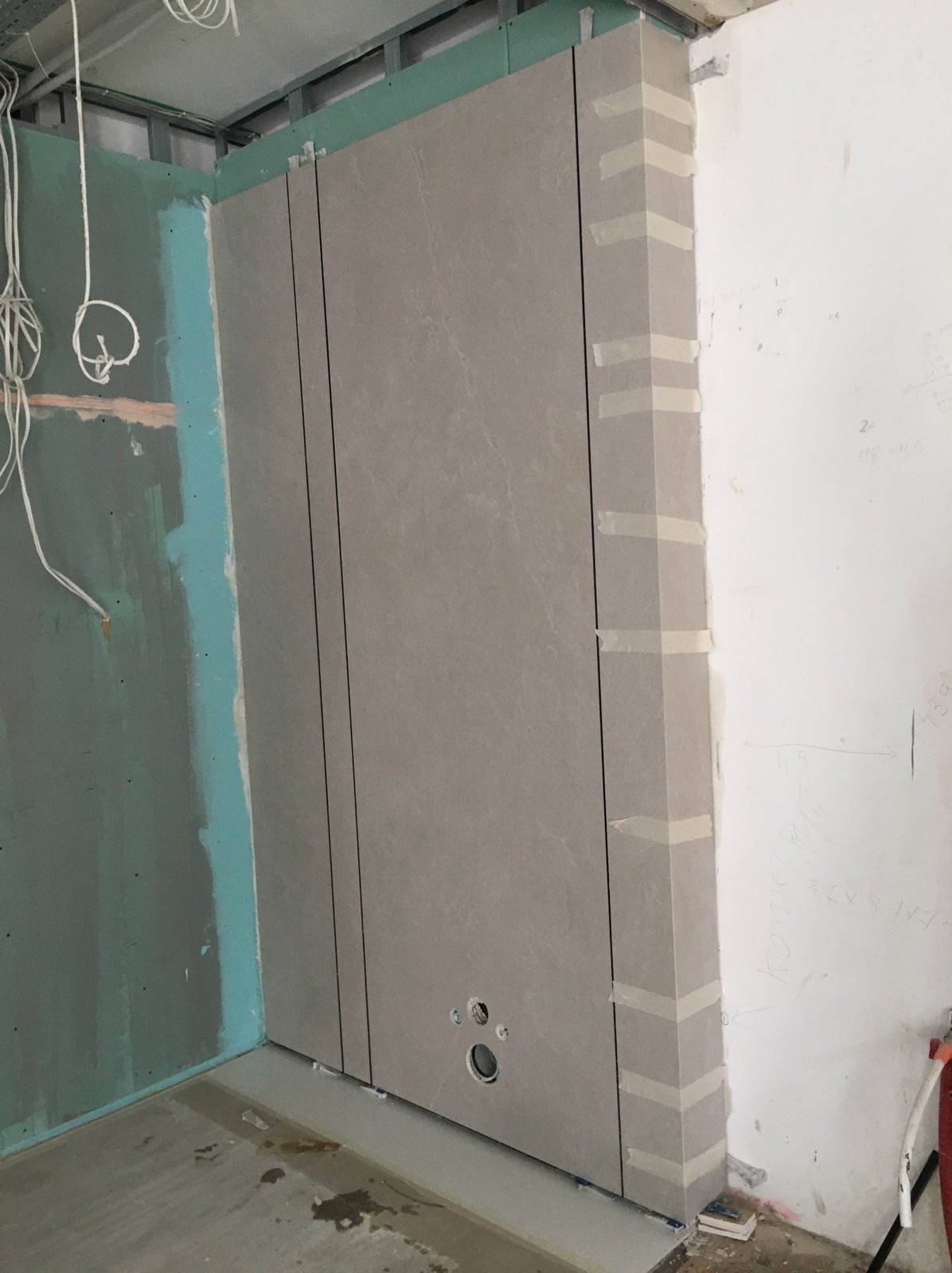 Ściana łazienki w trakcie montażu płyt wielkoformatowych, zabezpieczonych taśmą malarską na narożniku, widoczne otwory na instalacje wodne, surowy stan wykończenia ścian bocznych.