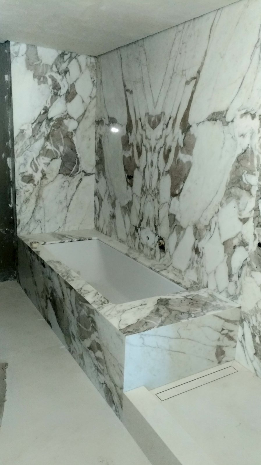 Biała wanna obudowana marmurem z szarymi żyłami, dopasowanym do okładziny ścian. Widoczny odpływ liniowy w podłodze i surowy fragment ściany z lewej strony.