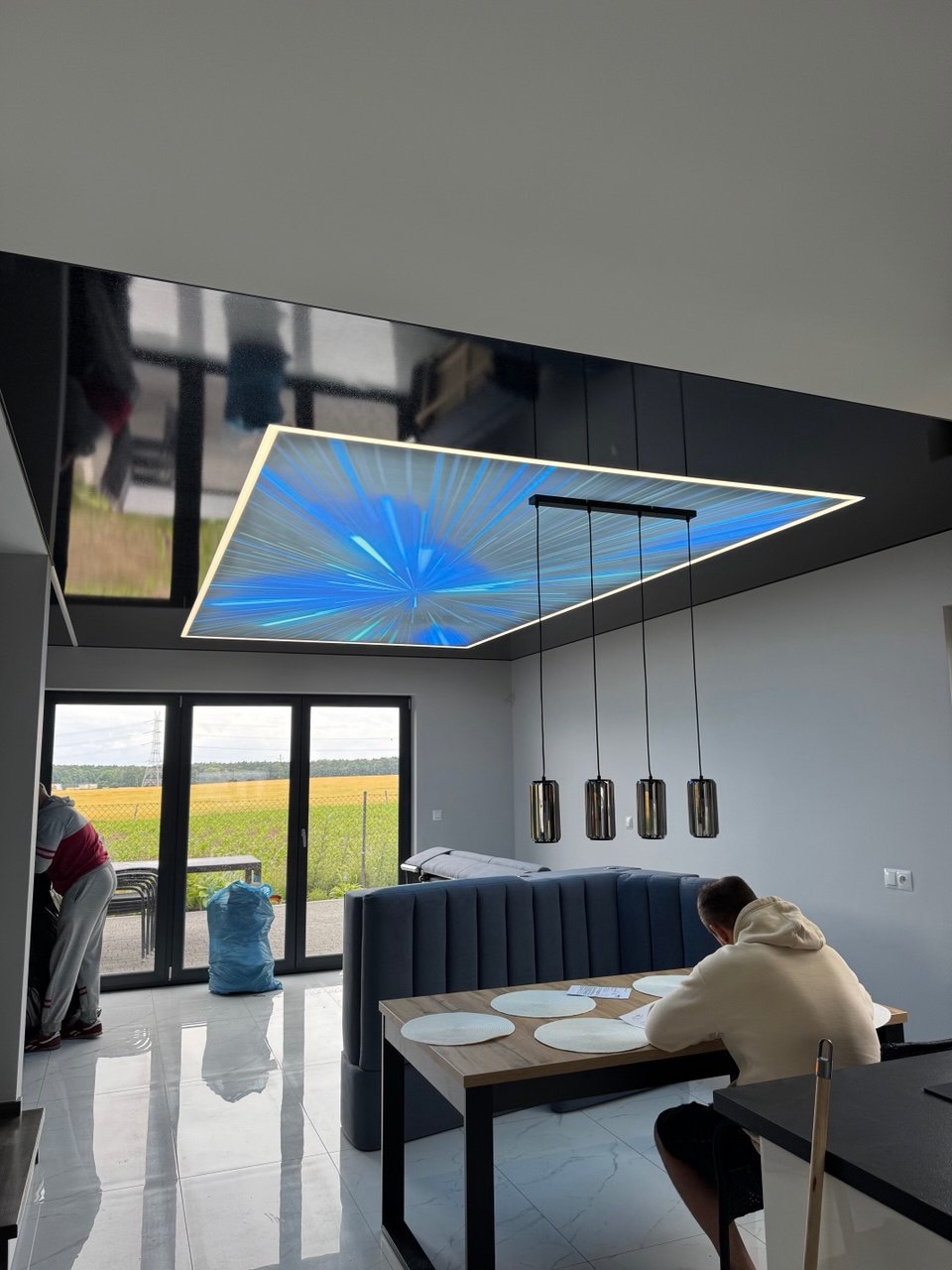 Nowoczesny salon z czarnym, błyszczącym sufitem i podświetlanym panelem z motywem rozgwieżdżonego nieba. Widoczne lampy wiszące i osoba siedząca przy stole.