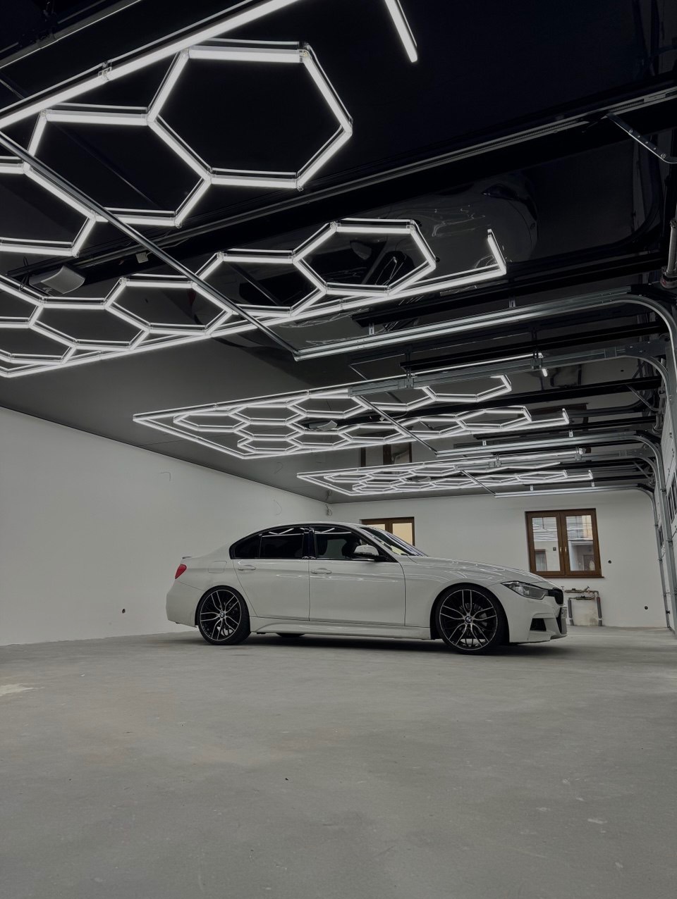 Biały samochód w garażu z czarnym, błyszczącym sufitem i heksagonalnym oświetleniem LED. Widoczny fragment bramy garażowej i okno w tle.