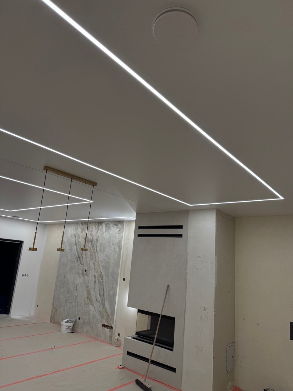 Nowoczesny salon w trakcie wykańczania, z liniowym oświetleniem LED na suficie, kominkiem i dekoracyjną ścianą. Widoczny etap prac wykończeniowych.