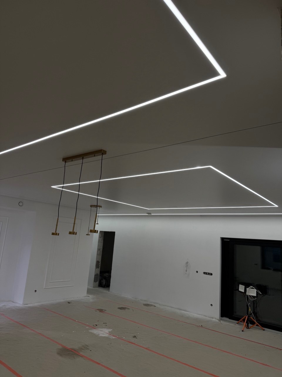 Nowoczesne oświetlenie sufitowe z liniami LED w kształcie prostokątów, uzupełnione wiszącymi lampami w minimalistycznym wnętrzu z surową podłogą.