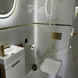 Luksusowa łazienka w Niemcach z białymi płytkami imitującymi marmur, złotymi akcentami i podświetlanym lustrem. Umywalka z szafką i wisząca toaleta.