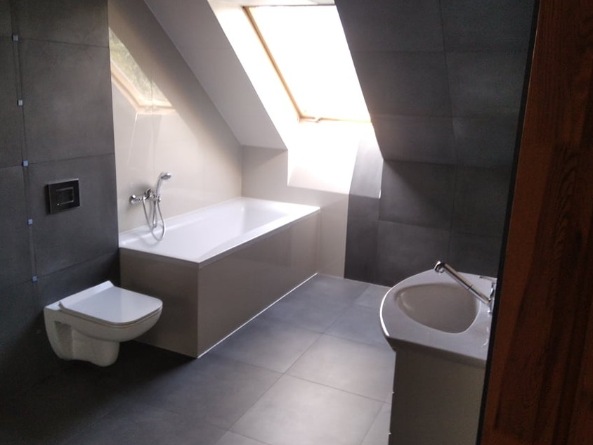 Nowoczesna łazienka na poddaszu z szarymi płytkami, wiszącą miską WC, białą wanną i umywalką, oświetlona naturalnym światłem z okna dachowego.