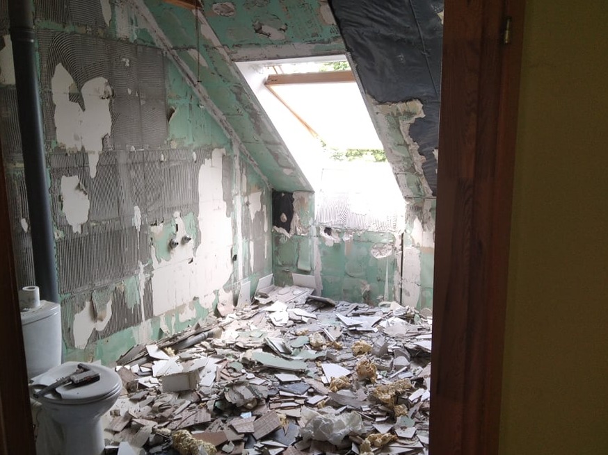 Łazienka na poddaszu w trakcie demontażu płytek, widoczne resztki kleju na ścianach, gruz na podłodze i narzędzia na sedesie, okno dachowe w tle.