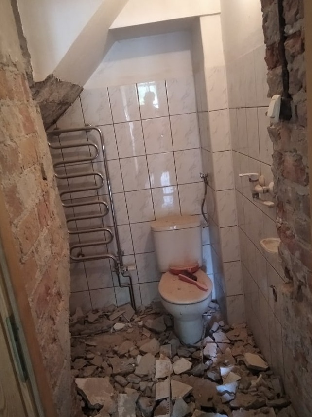 Łazienka w trakcie demontażu płytek, widoczne gruzowisko na podłodze, odsłonięte cegły na ścianie, wiszący grzejnik drabinkowy i toaleta z brudną deską.