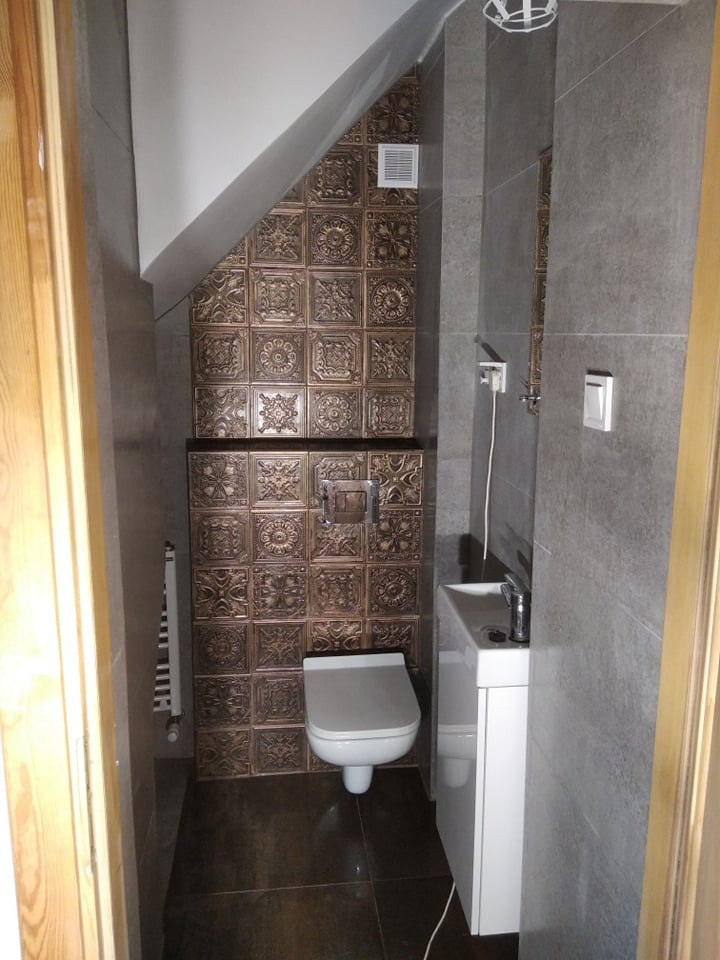 Mała łazienka na poddaszu z toaletą podwieszaną, umywalką z szafką i dekoracyjnymi, brązowymi płytkami w strefie WC.