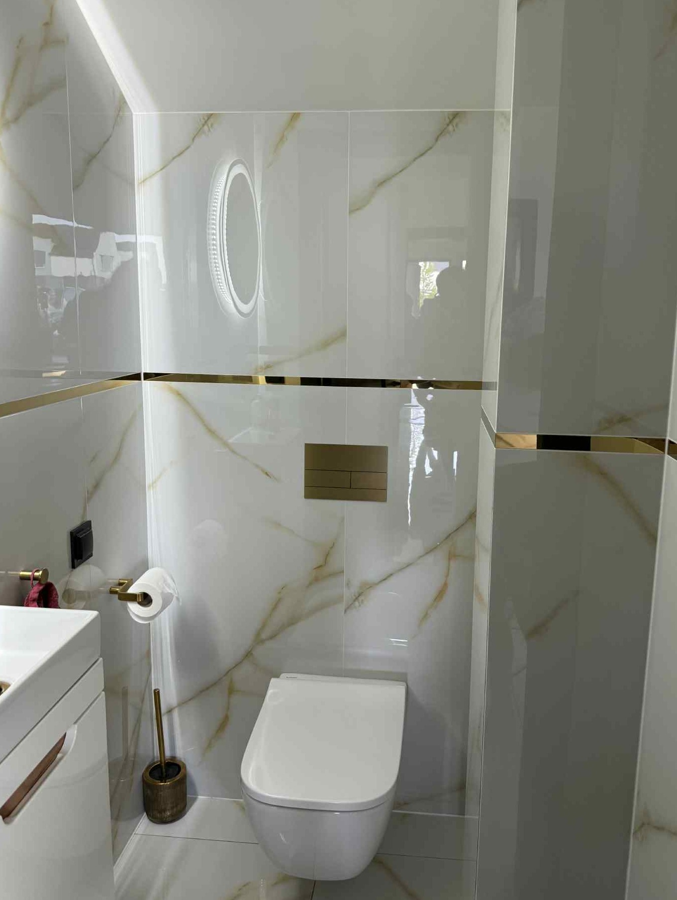 Elegancka toaleta z białymi płytkami imitującymi marmur, złotymi akcentami i podświetlanym lustrem. Widoczna toaleta podwieszana i szczotka do WC w złotym kolorze.