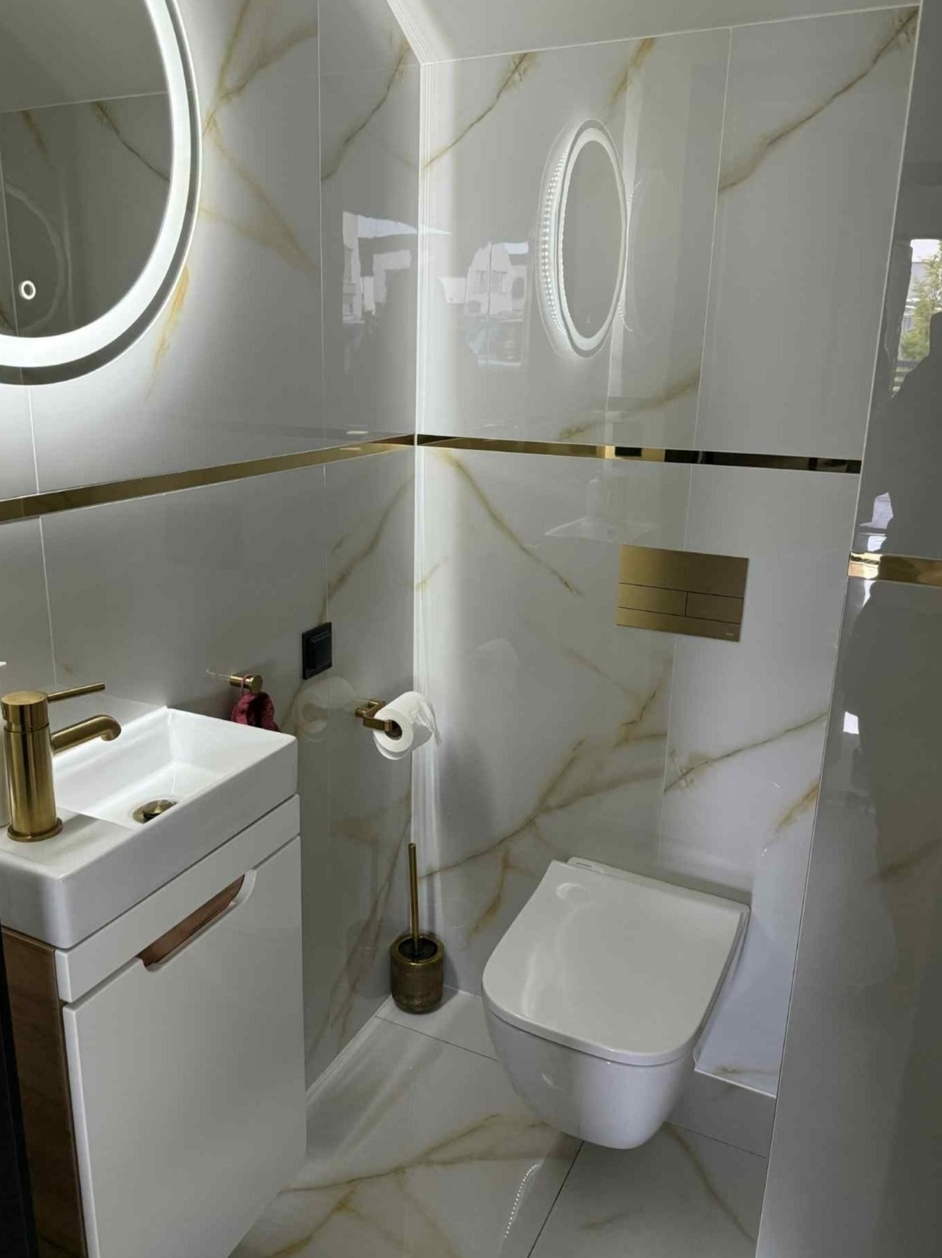 Luksusowa łazienka w Niemcach z białymi płytkami imitującymi marmur, złotymi akcentami i podświetlanym lustrem. Umywalka z szafką i wisząca toaleta.