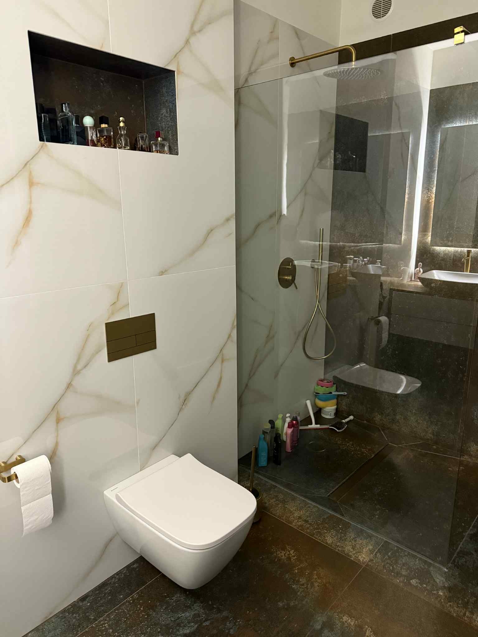 Elegancka łazienka z toaletą podwieszaną, kabiną prysznicową typu walk-in i marmurowymi płytkami z żyłkowaniem w odcieniach złota. Widoczna wnęka z kosmetykami.