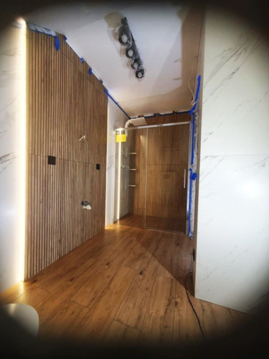 Nowoczesna łazienka w trakcie wykończenia: drewniane panele ścienne z oświetleniem LED, kabina prysznicowa ze szklanymi drzwiami i drewnopodobna podłoga.