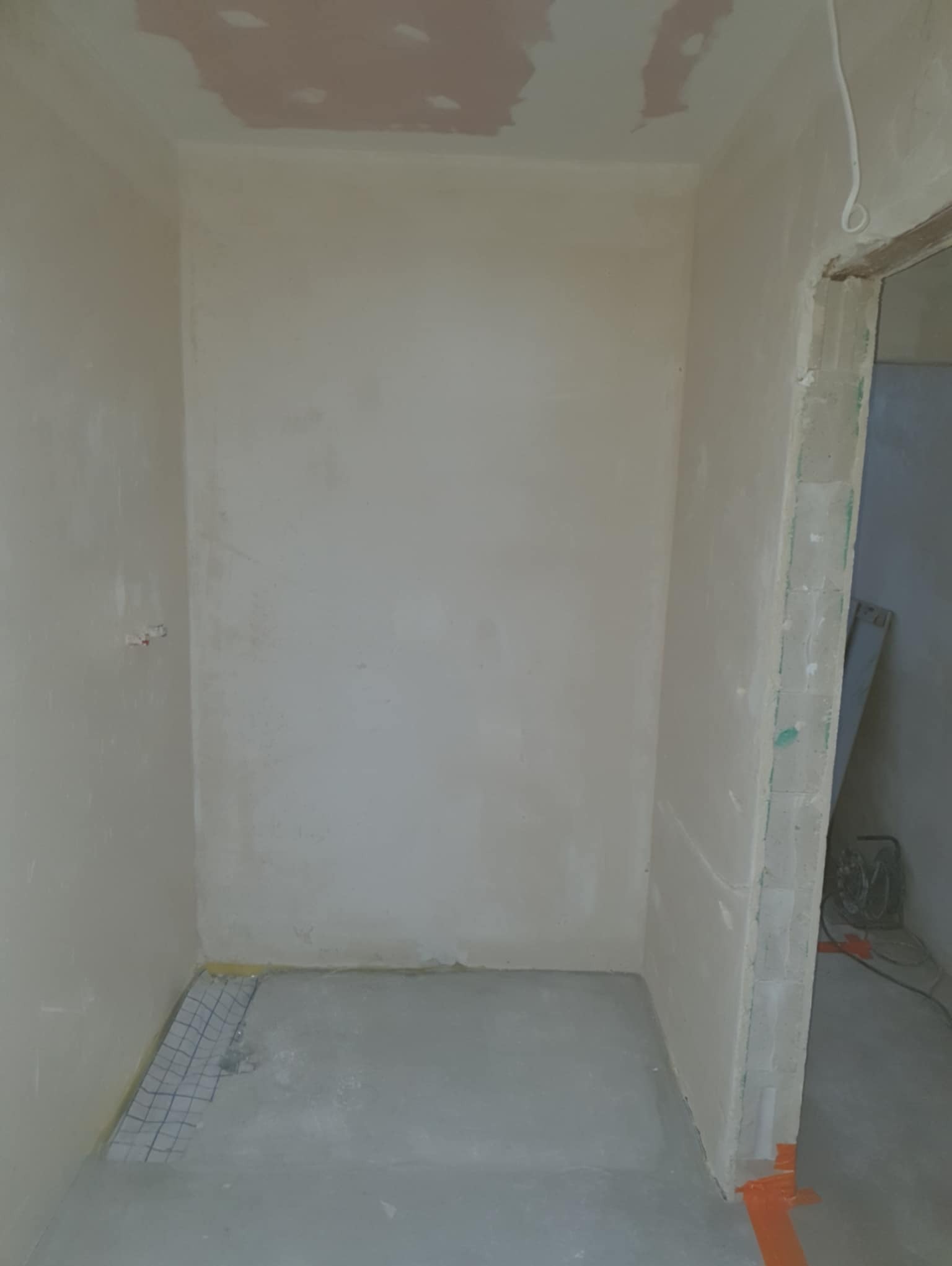 Surowa łazienka w trakcie remontu: ściany po gładzi, fragment siatki pod ogrzewanie podłogowe, widoczne ślady po łatach na suficie. Minimalistyczny kadr.