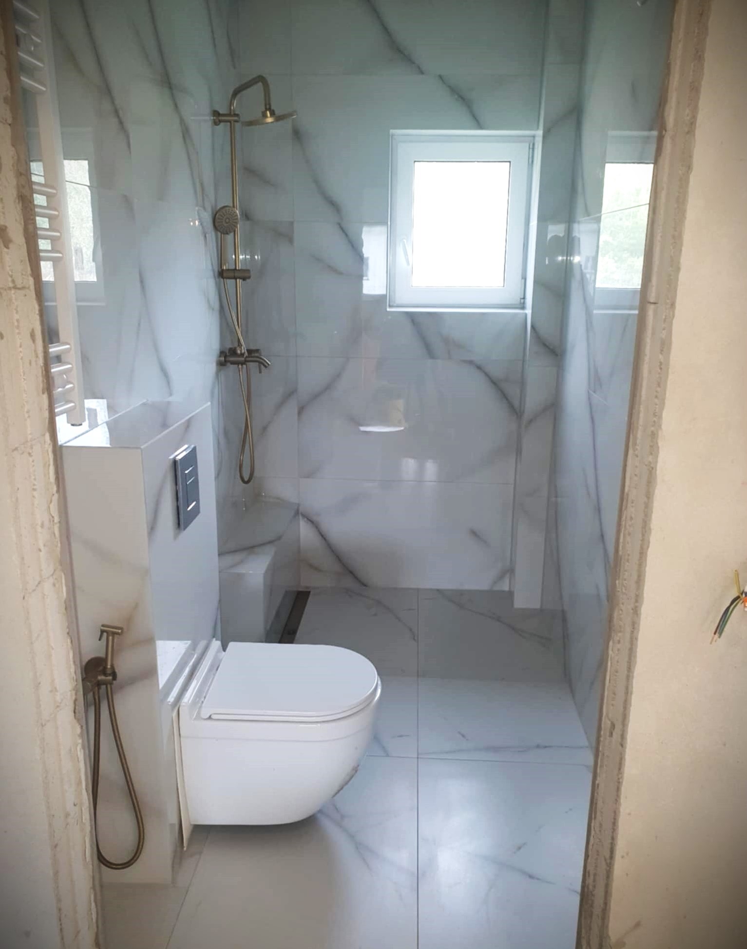 Wykończona łazienka z marmurowymi płytkami, podwieszaną toaletą z bidetką w kolorze starego złota i deszczownicą. Widoczne okno i odpływ liniowy.